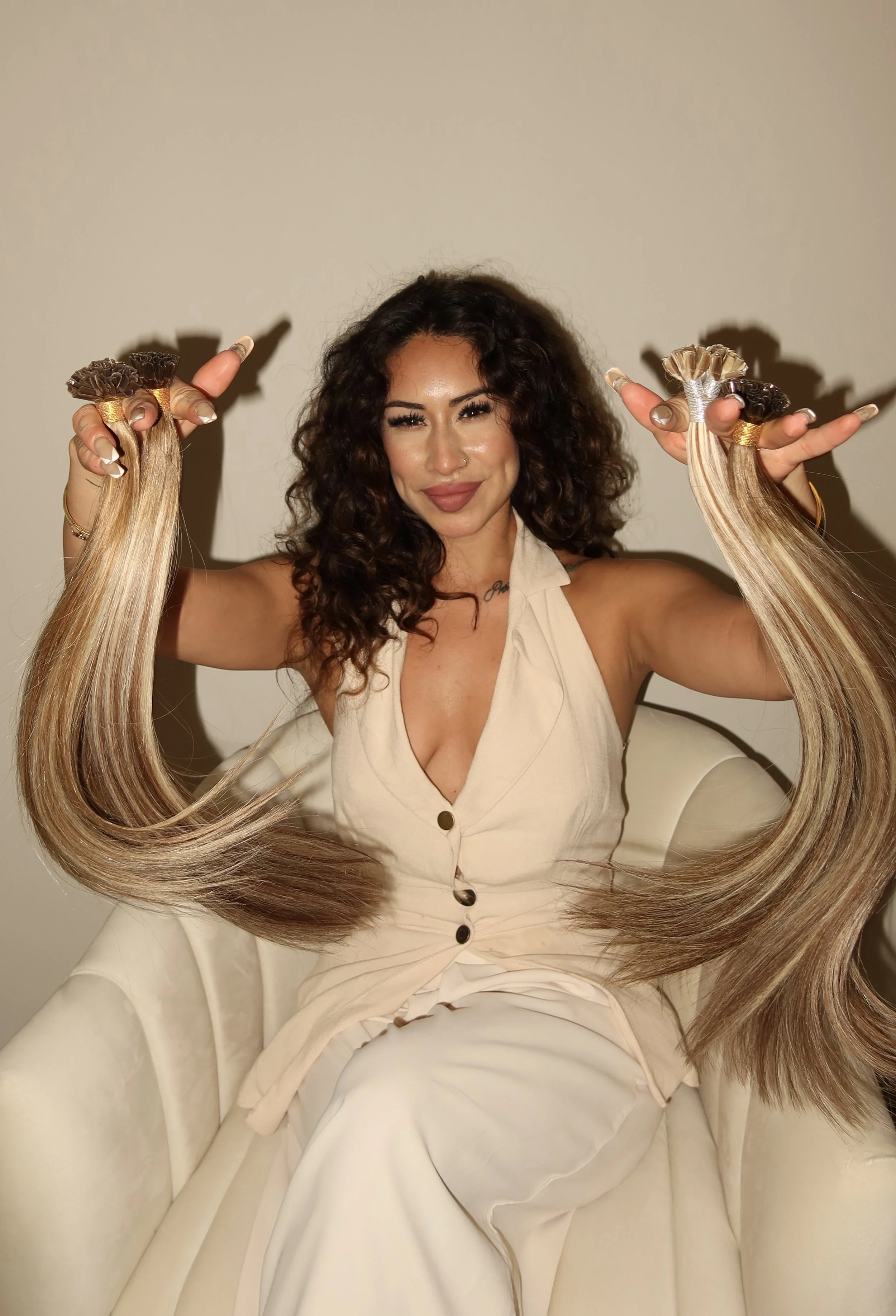 A woman with curly hair holding strands of her long blonde hair in each hand, sitting on a beige chair with a plain, light-colored background.