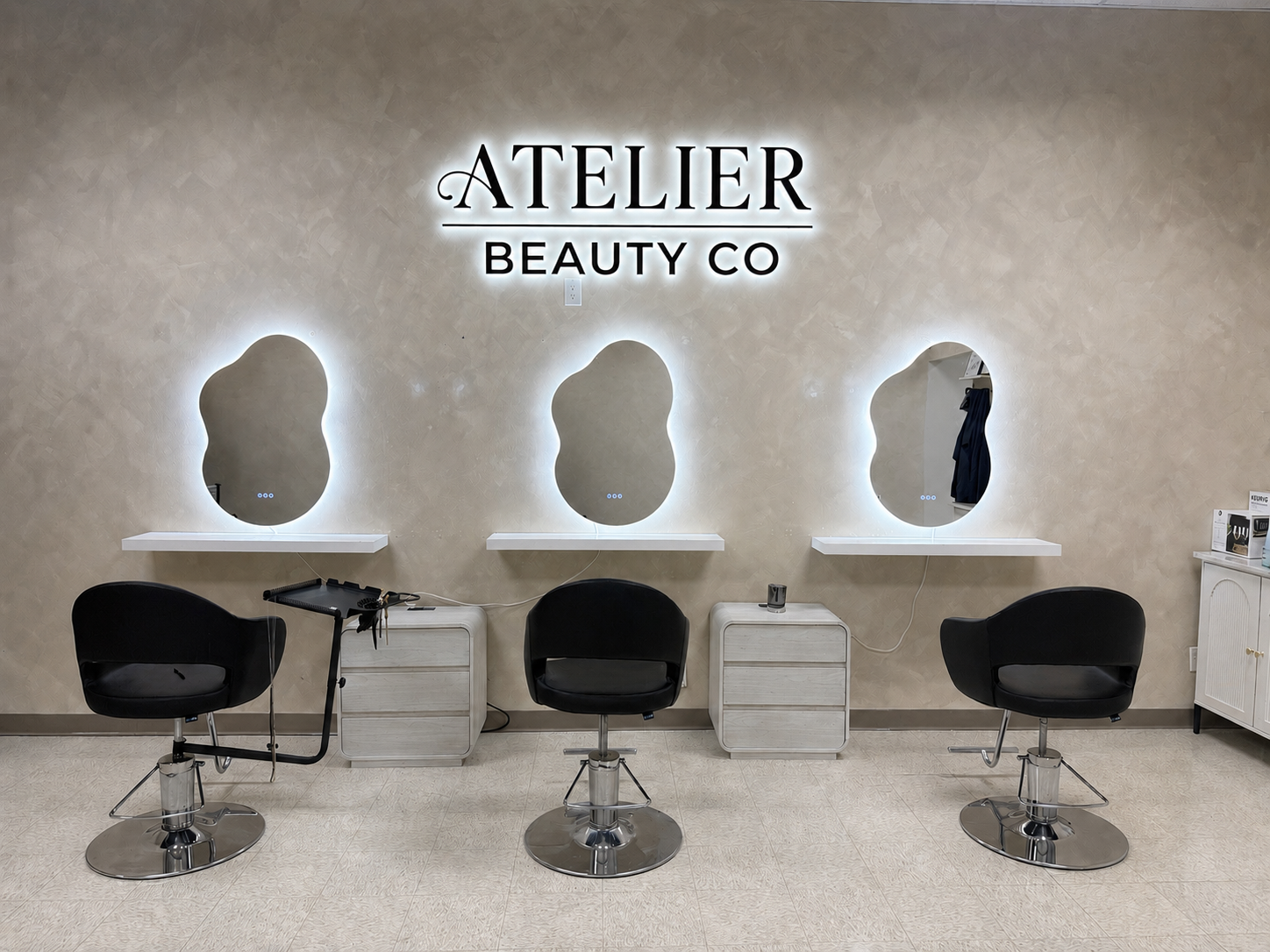 A beauty salon with three black chairs, three mirrors with backlit outlines, and a beige textured wall with a sign that reads 'ATELIER BEAUTY CO'.