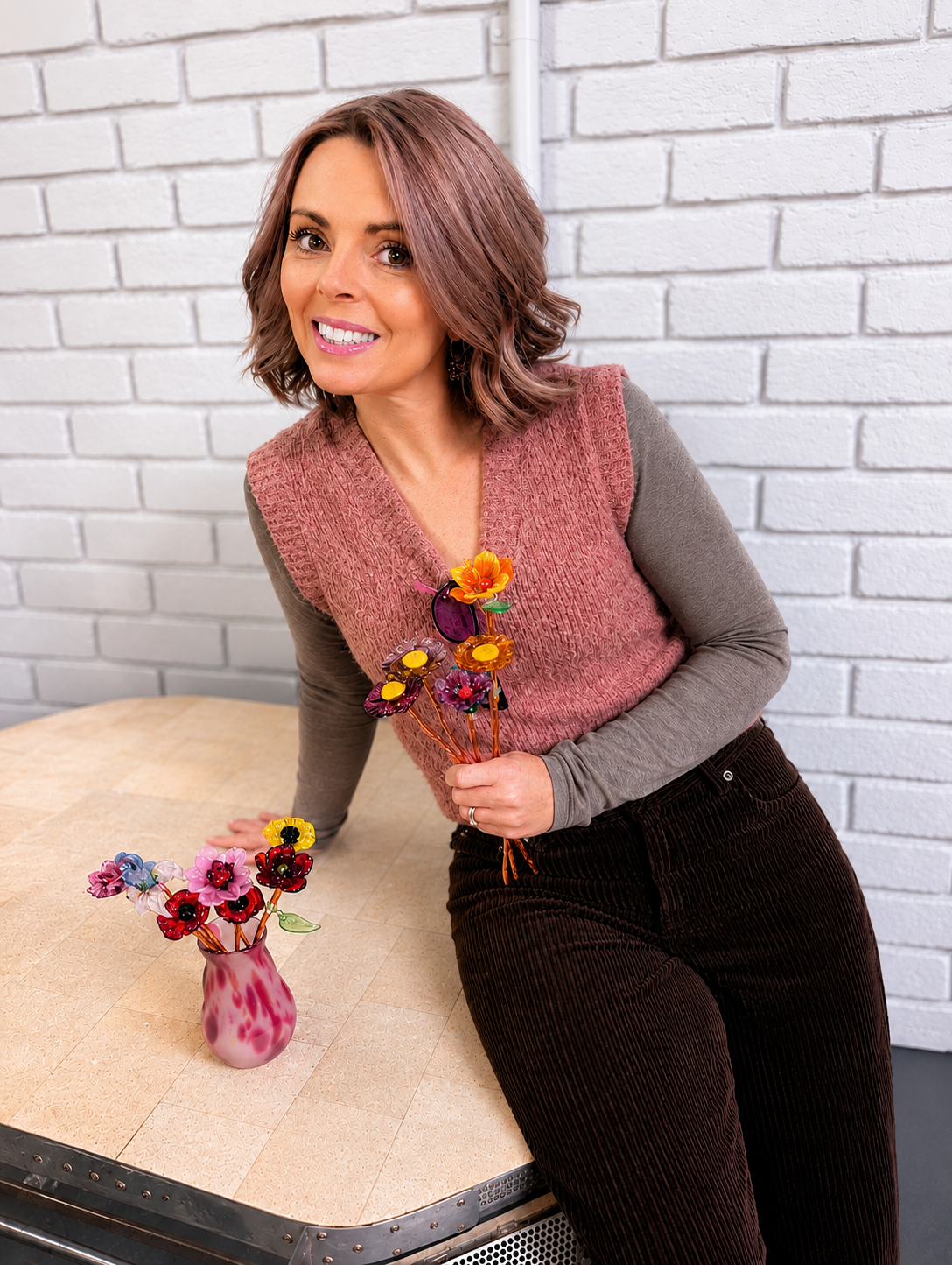 A woman with shoulder-length wavy brown hair and a pinkish-brown top, standing next to a table with a pink and white vase filled with colorful artificial flowers, against a white brick wall.