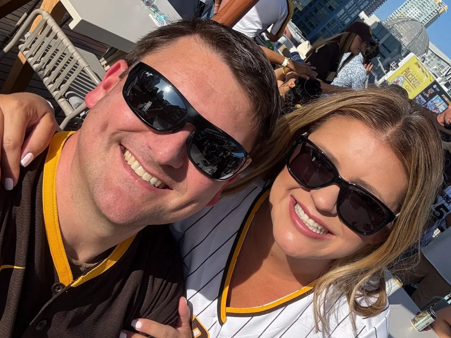 Two smiling people wearing sunglasses taking a selfie at an outdoor event on a rooftop during sunny weather with city buildings in the background.