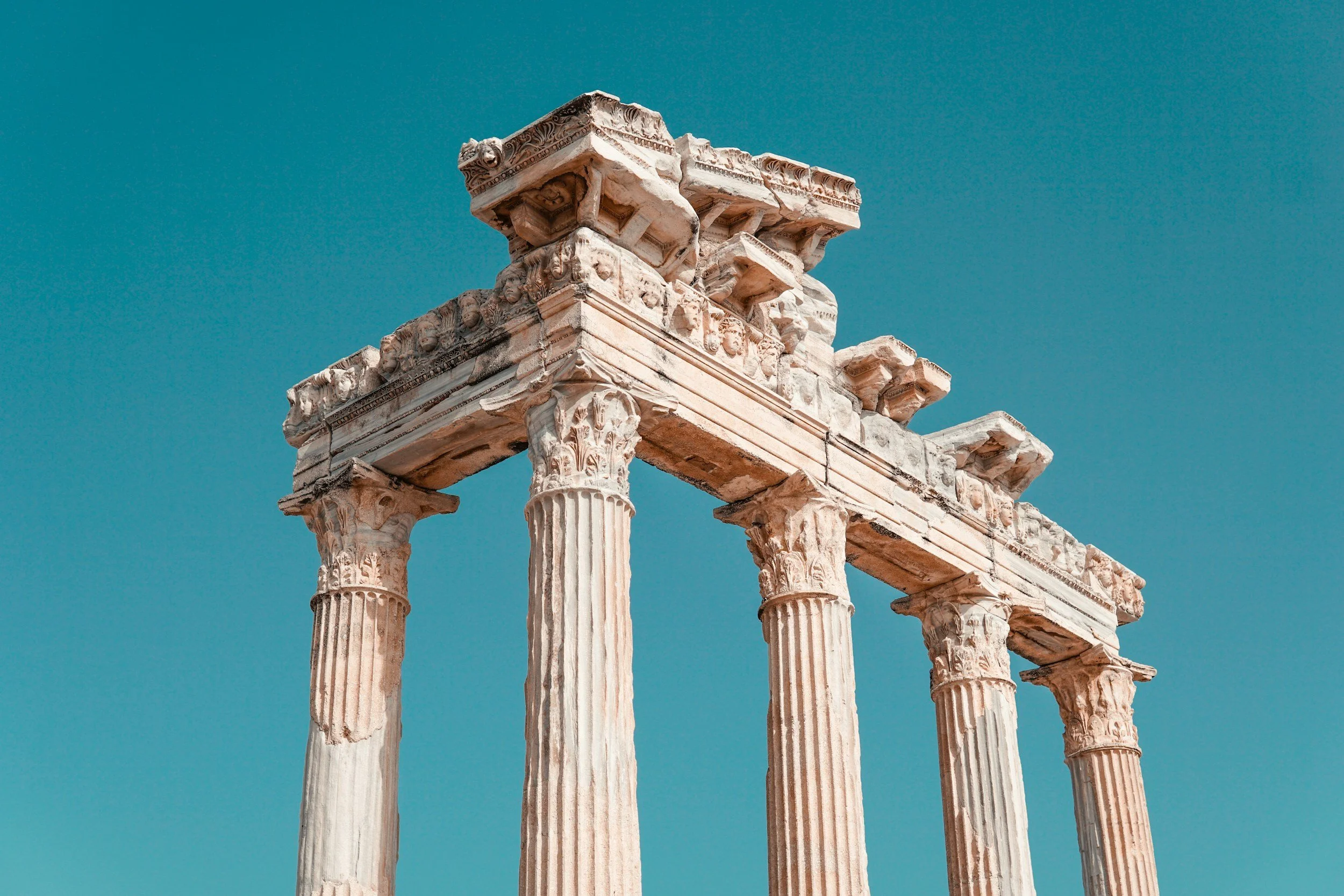 Ancient Greek ruins with six tall, fluted columns and decorated capitals against a clear blue sky.