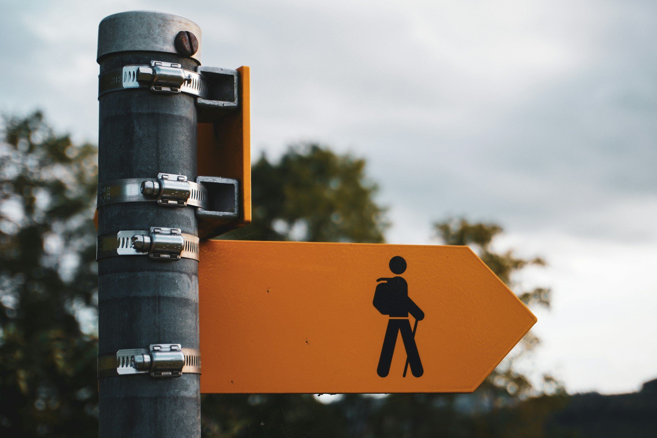 Close-up of a metal pole with multiple hose clamps and an orange arrow sign indicating pedestrian walking route against a cloudy sky and blurred trees in the background.