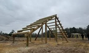 Construction of a wooden outdoor structure with beams and supports on a dirt field under a cloudy sky.