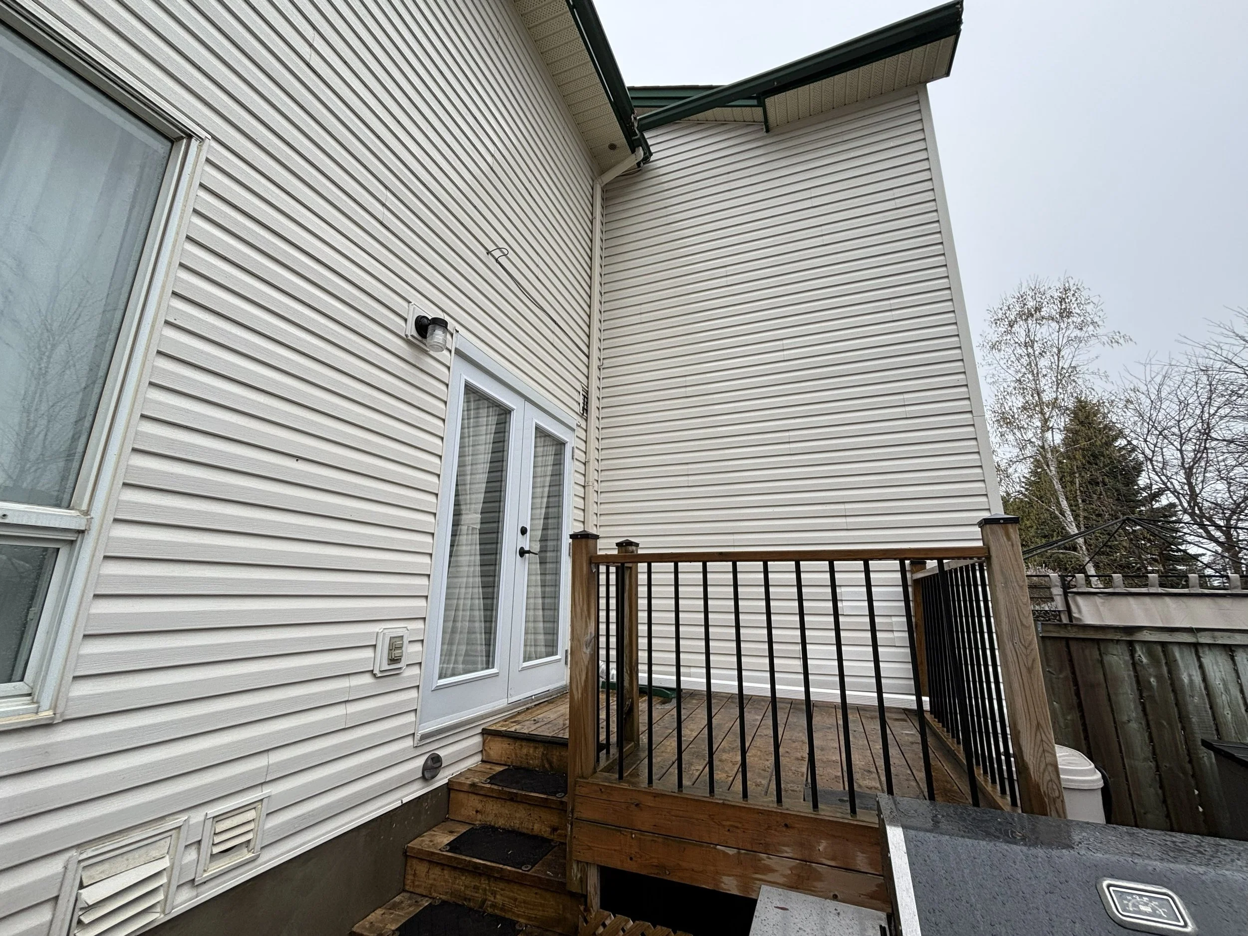 Backyard deck attached to a beige house with siding, with stairs leading down, black railing, and a wooden railing. The sky is overcast and part of a tree is visible on the right.