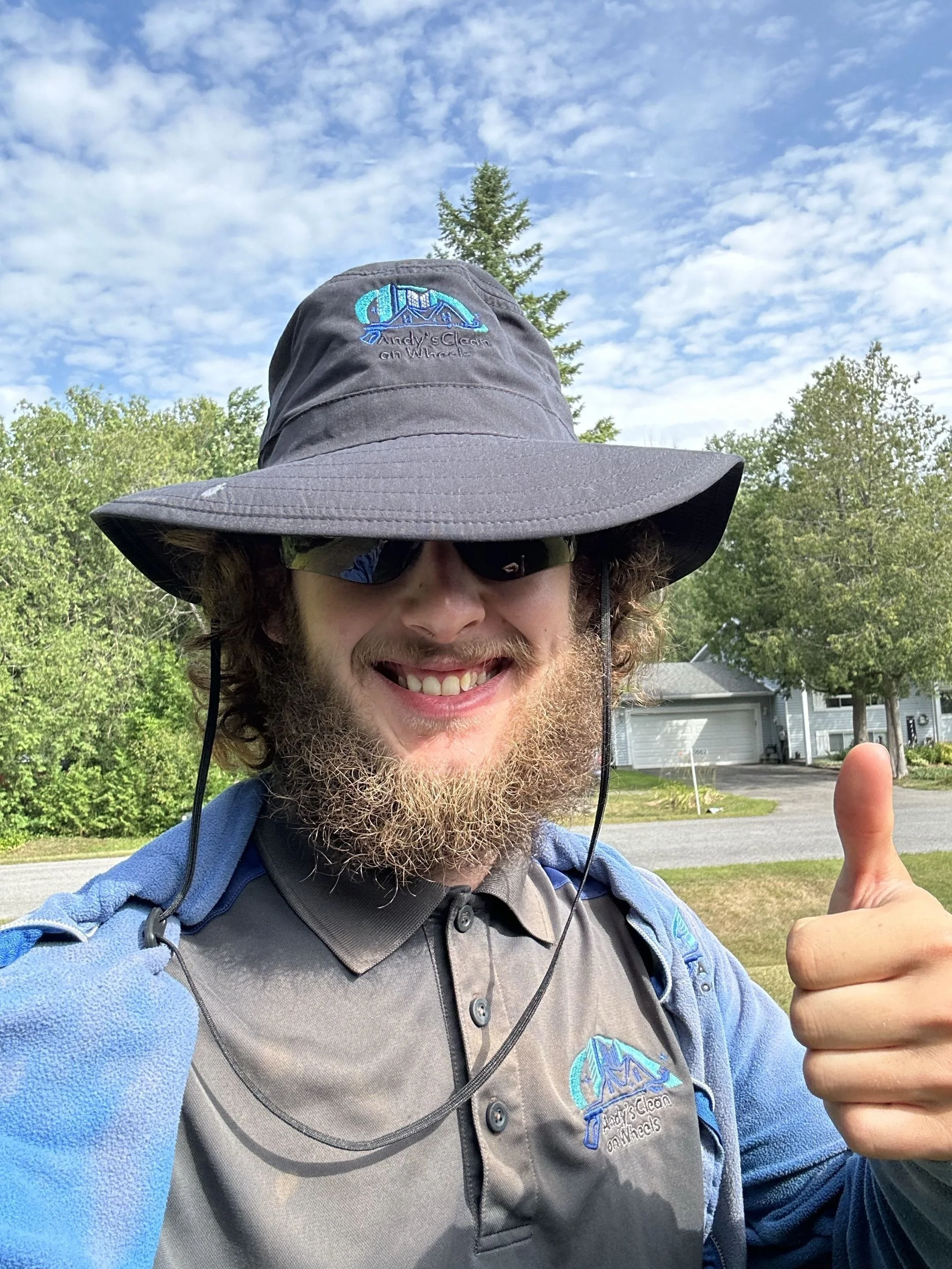 A man smiling outdoors wearing a wide-brimmed hat with an embroidered logo and text, sunglasses, and a blue jacket giving a thumbs-up. There are trees and a house in the background.