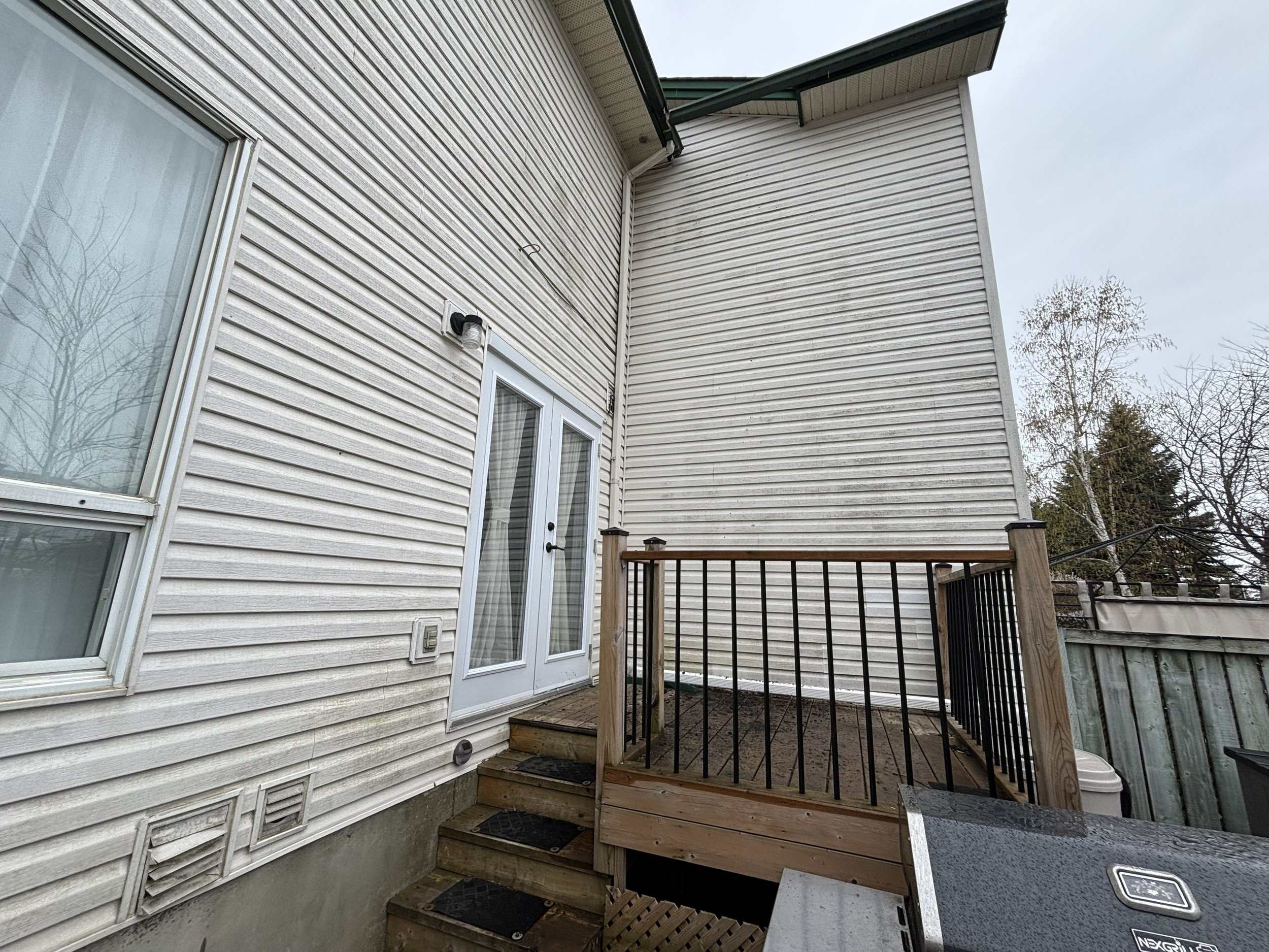 A small wooden deck with stairs and black metal railing attached to the back of a beige house with vinyl siding. There is a sliding glass door with a white frame leading into the house. A black outdoor light fixture is mounted on the house near the door. The sky is overcast, and there are leafless trees visible in the background.