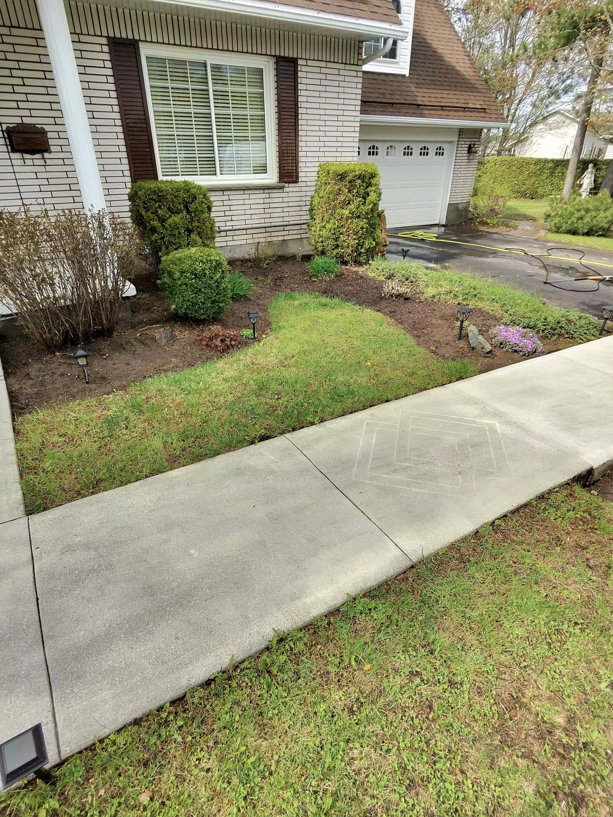 Front yard of house with a concrete sidewalk, green grass, and garden beds with plants and bushes. Part of a white brick house with white garage door and brown window shutters is visible.