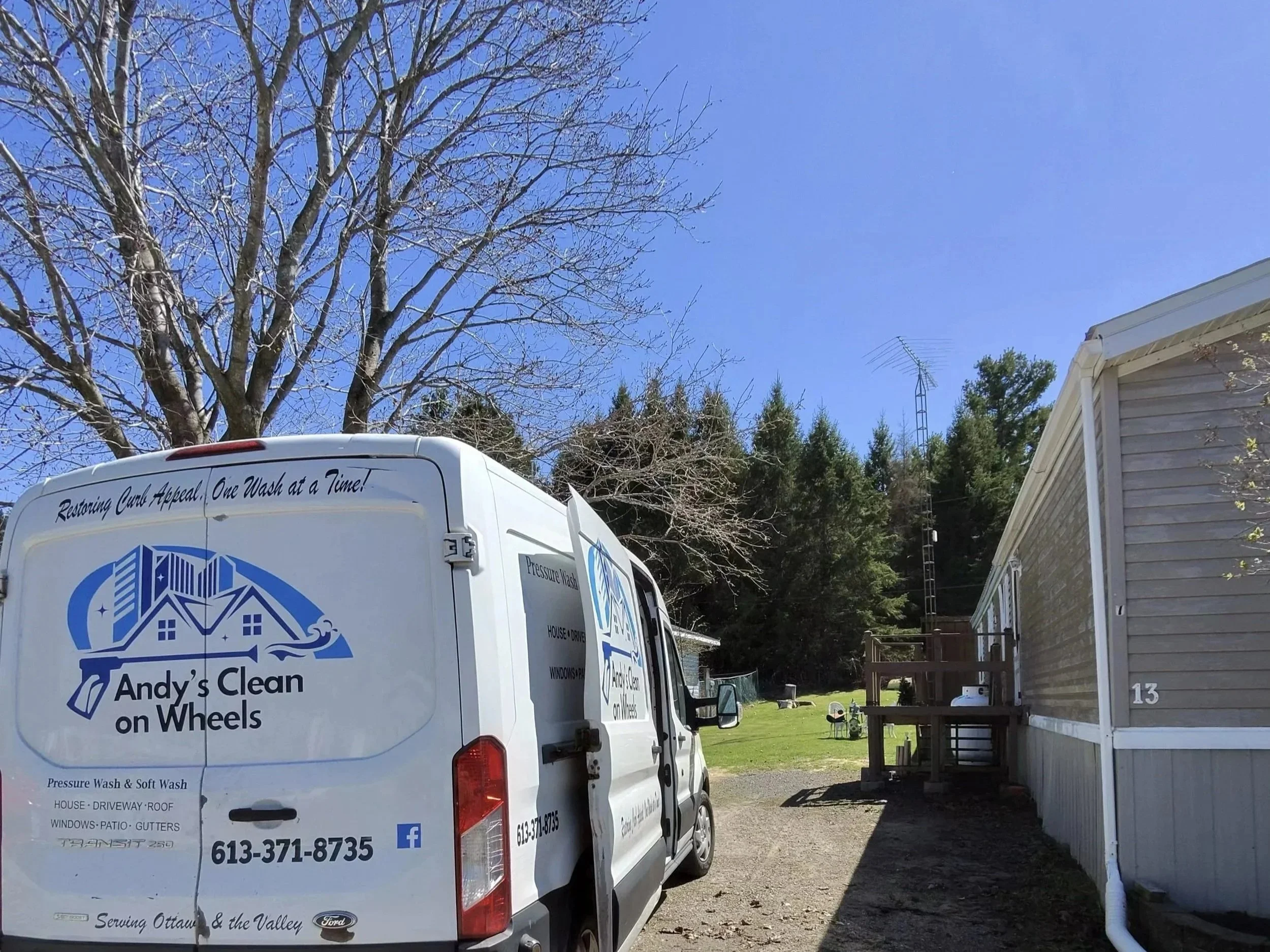 A white service van with the logo and contact information of Andy's Clean on Wheels parked in a residential driveway. The background features a leafless tree, a house with a deck, and a clear blue sky.