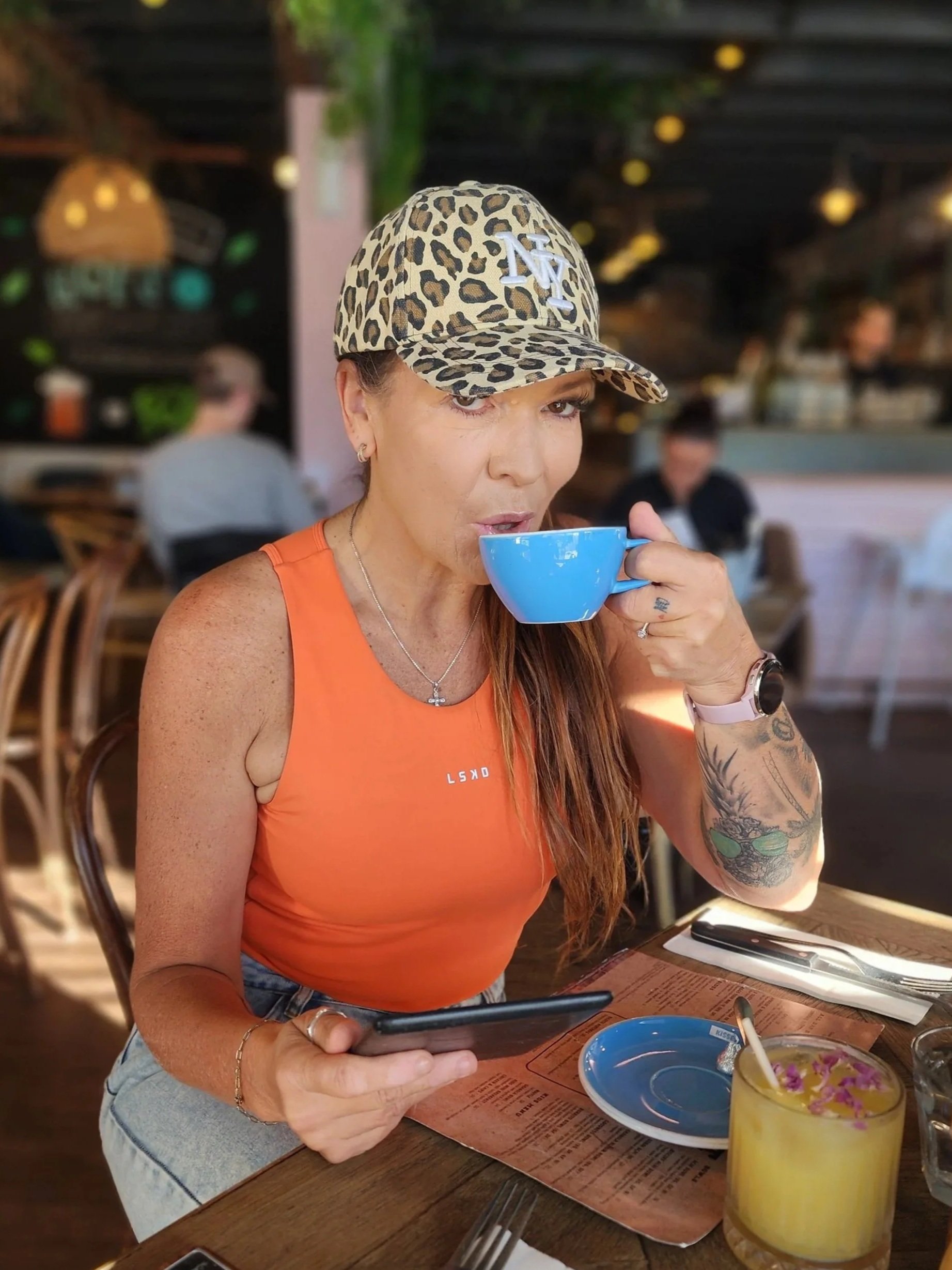 Woman with long red hair wearing an orange tank top and leopard-print baseball cap sitting at a restaurant table, sipping from a blue cup. There is a yellow cocktail with a straw and flowers on the garnish, a black smartphone, and a red placemat with a menu on the table. Blurred background of other diners and hanging lights.