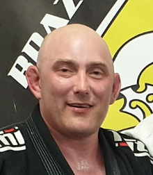 A bald man in a black martial arts gi standing in front of a yellow and black background.