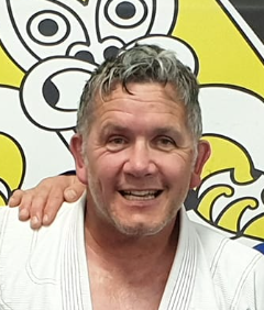A smiling man with short, curly gray hair wearing a white martial arts gi, standing in front of a yellow and black cartoon bee background.