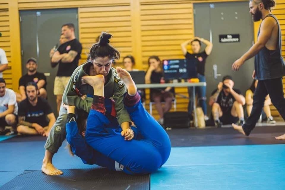 Two women competing in Brazilian Jiu-Jitsu on a blue mat, with one woman in a green gi controlling the other in a blue gi; spectators and a referee are watching.