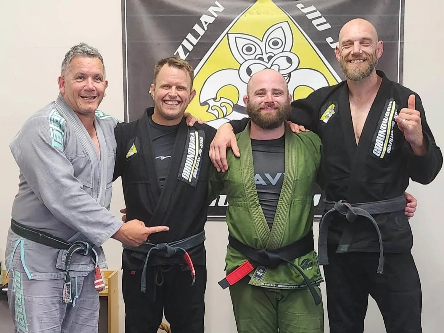 Four men in Brazilian Jiu-Jitsu gis standing together, smiling, with one man pointing at the person next to him. They are in a martial arts dojo with a banner featuring a tribal mask design in the background.