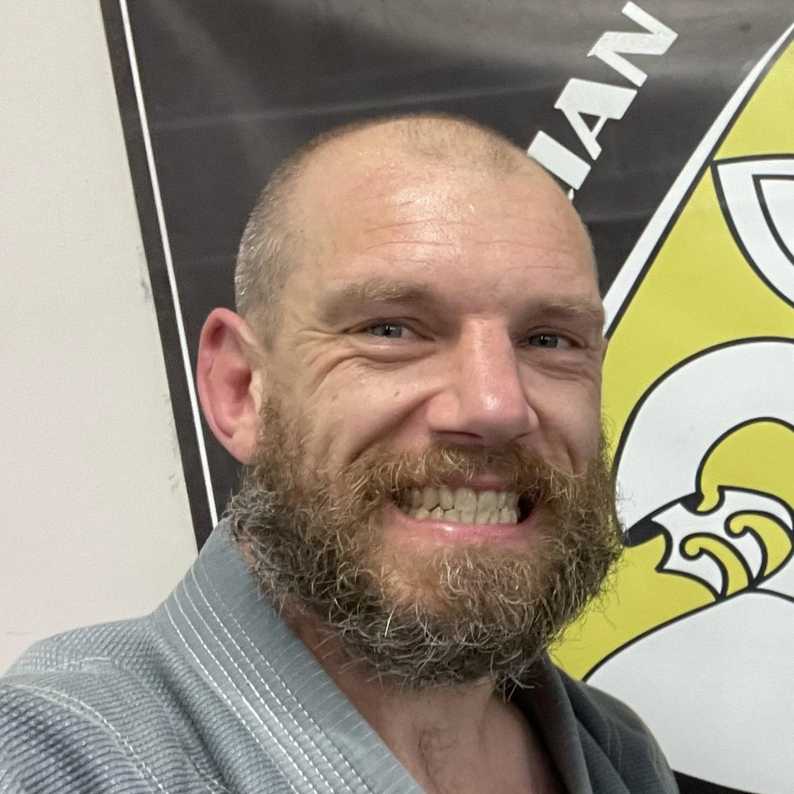A man with a beard and short hair smiling, wearing a gray suit jacket, in front of a black and yellow banner with partial text visible.