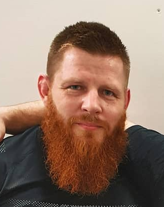 A man with short brown hair, a full reddish beard, and blue eyes, wearing a dark shirt, poses with his hand resting on his head against a plain light-colored background.