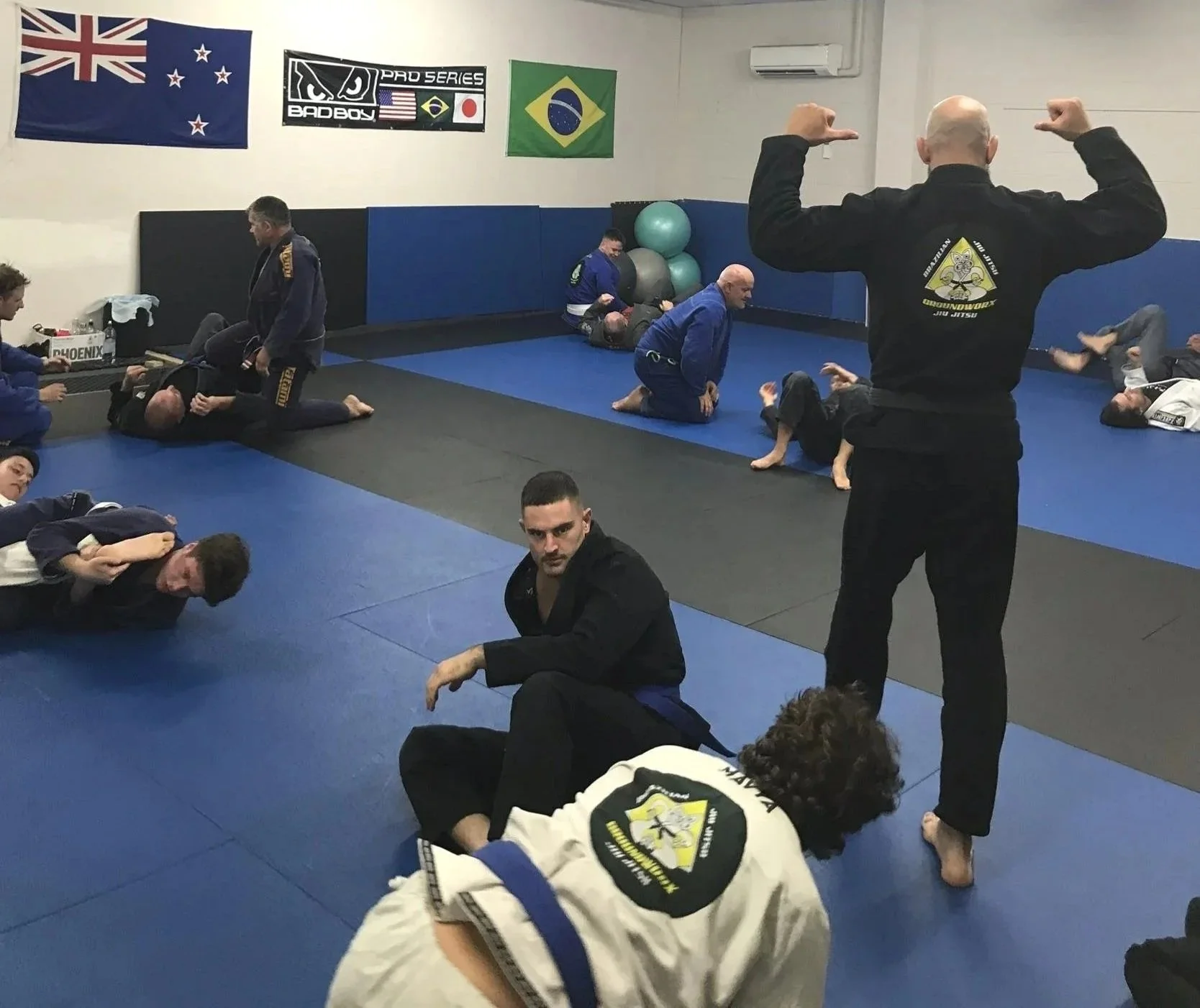 Brazilian Jiu-Jitsu class with students practicing on mats, instructor overseeing, three flags on wall including New Zealand, Brazil, and another with a logo, exercise balls in corner.
