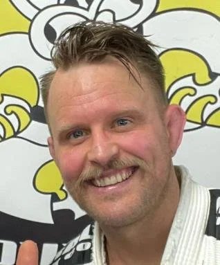 A smiling man with short brown hair and a mustache, wearing a white shirt with black stripes, standing in front of a cartoon tiger mural.