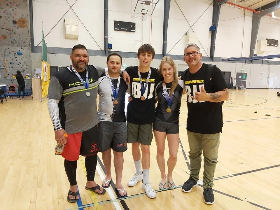 A group of five people standing together in a gymnasium, wearing medals and sportswear, celebrating a sports achievement.