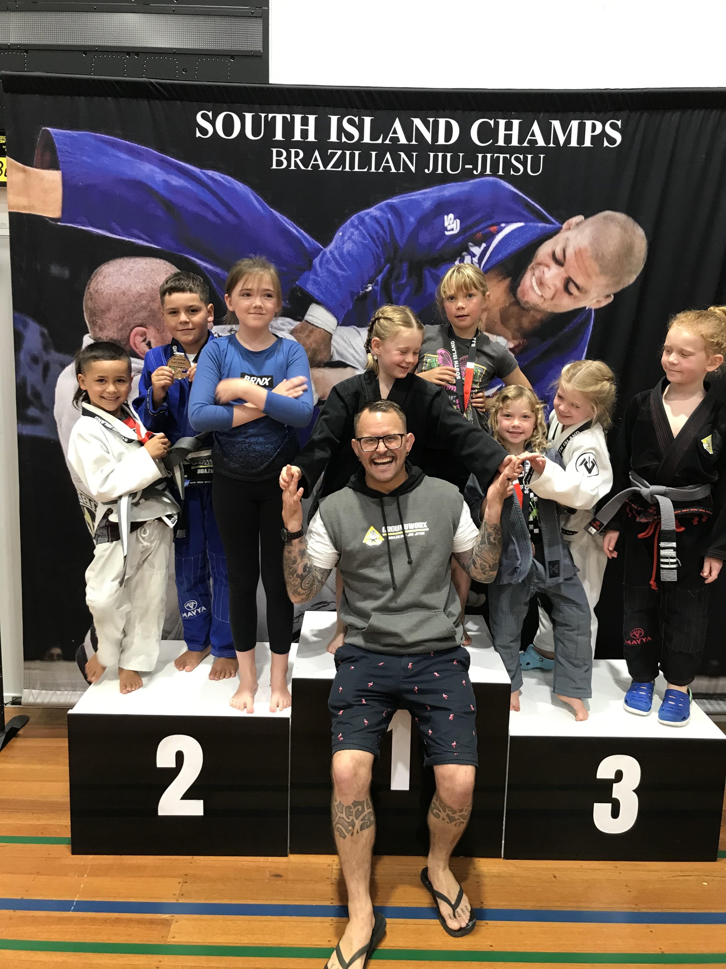 Group of children in Brazilian Jiu-Jitsu uniforms standing on a winners' podium with medals, in front of a large photo of a Brazilian Jiu-Jitsu athlete in blue gi on a black backdrop that reads 'South Island Champs Brazilian Jiu-Jitsu'.