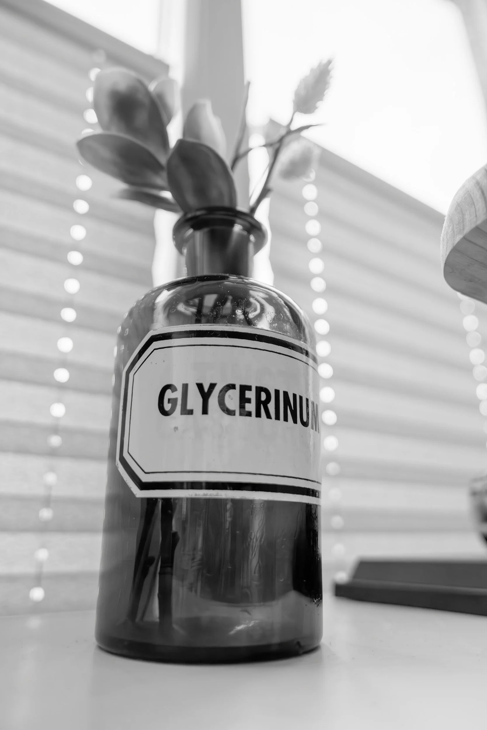 A plastic Glycerin bottle with a plant inside, placed on a white surface with horizontal blinds in the background.
