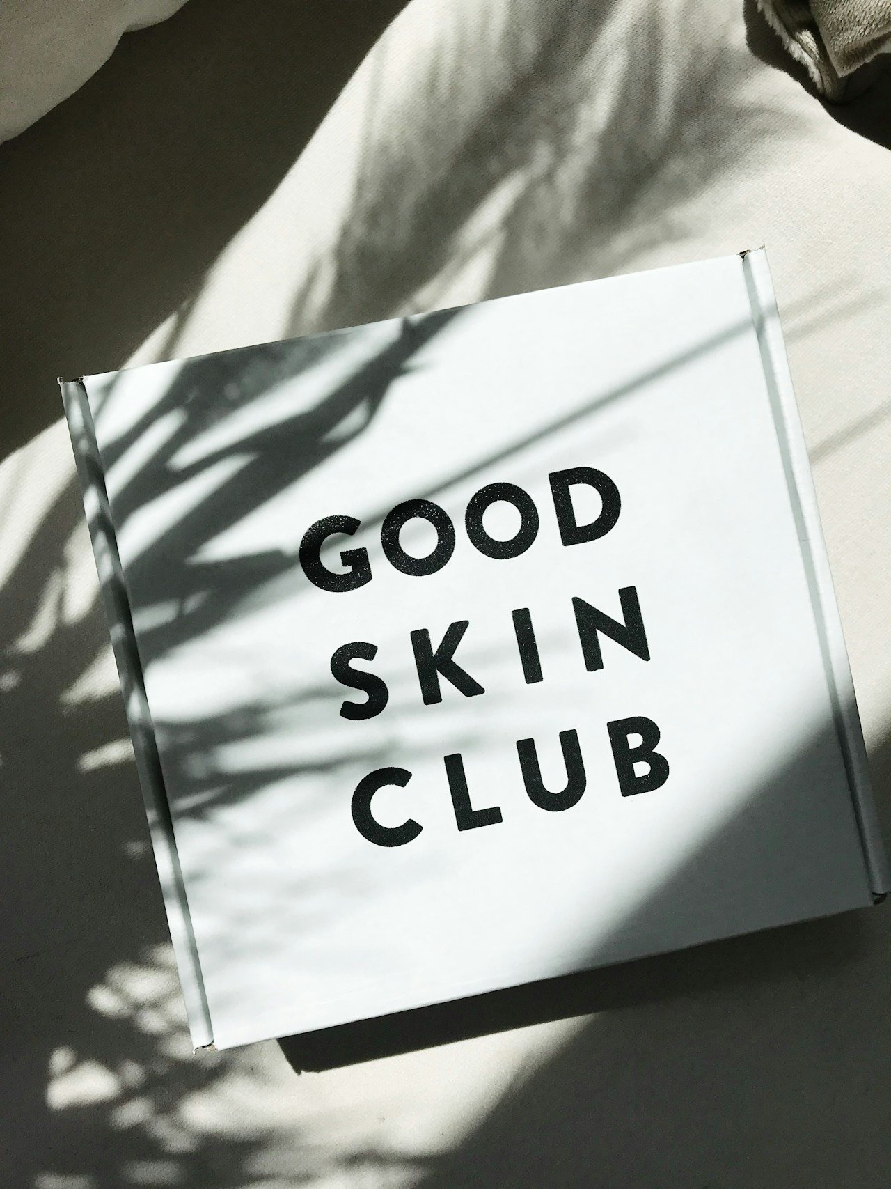 A white box with black text reading 'Good Skin Club' on the top, with shadows of leaves cast across it, placed on a light surface.