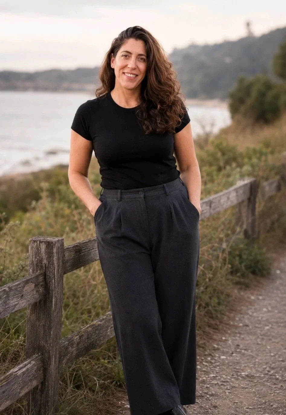 Jennifer Vulgan, smiling and standing outdoors by a wooden fence with water and hills in the background.
