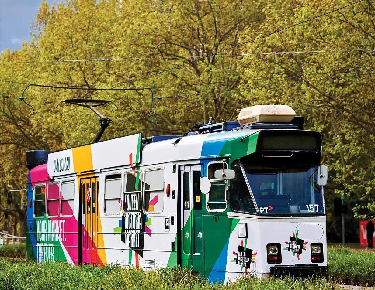 Colorful tram decorated with vibrant geometric patterns advertising Queen Victoria Market in Melbourne, with trees and a blue sky in the background.