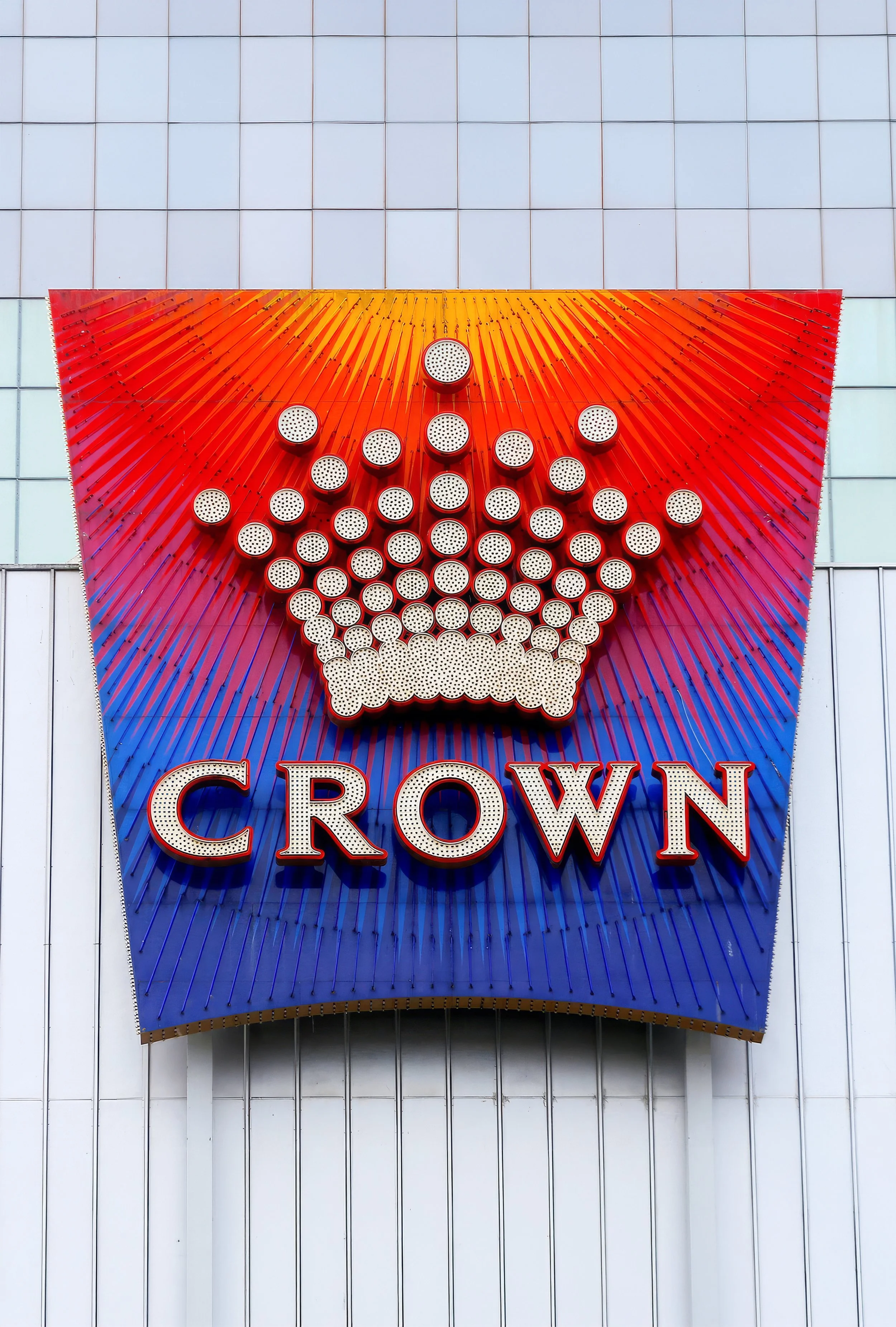 A large vertical sign with the word "CROWN" at the bottom, featuring a stylized crown design made of metal tubes on a gradient background of red and blue rays.
