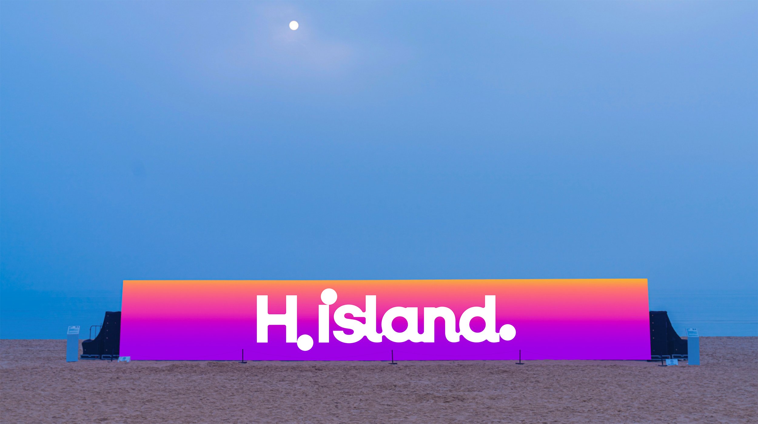 A large colorful sign on the beach reads 'H!land'. The sky above is clear with a visible moon.