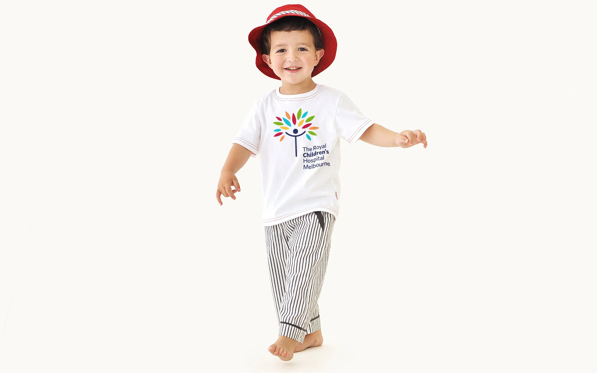 A young boy wearing a red hat, a white T-shirt with a colorful logo from The Royal Children's Hospital Melbourne, and black-and-white striped pants, smiling and walking barefoot.