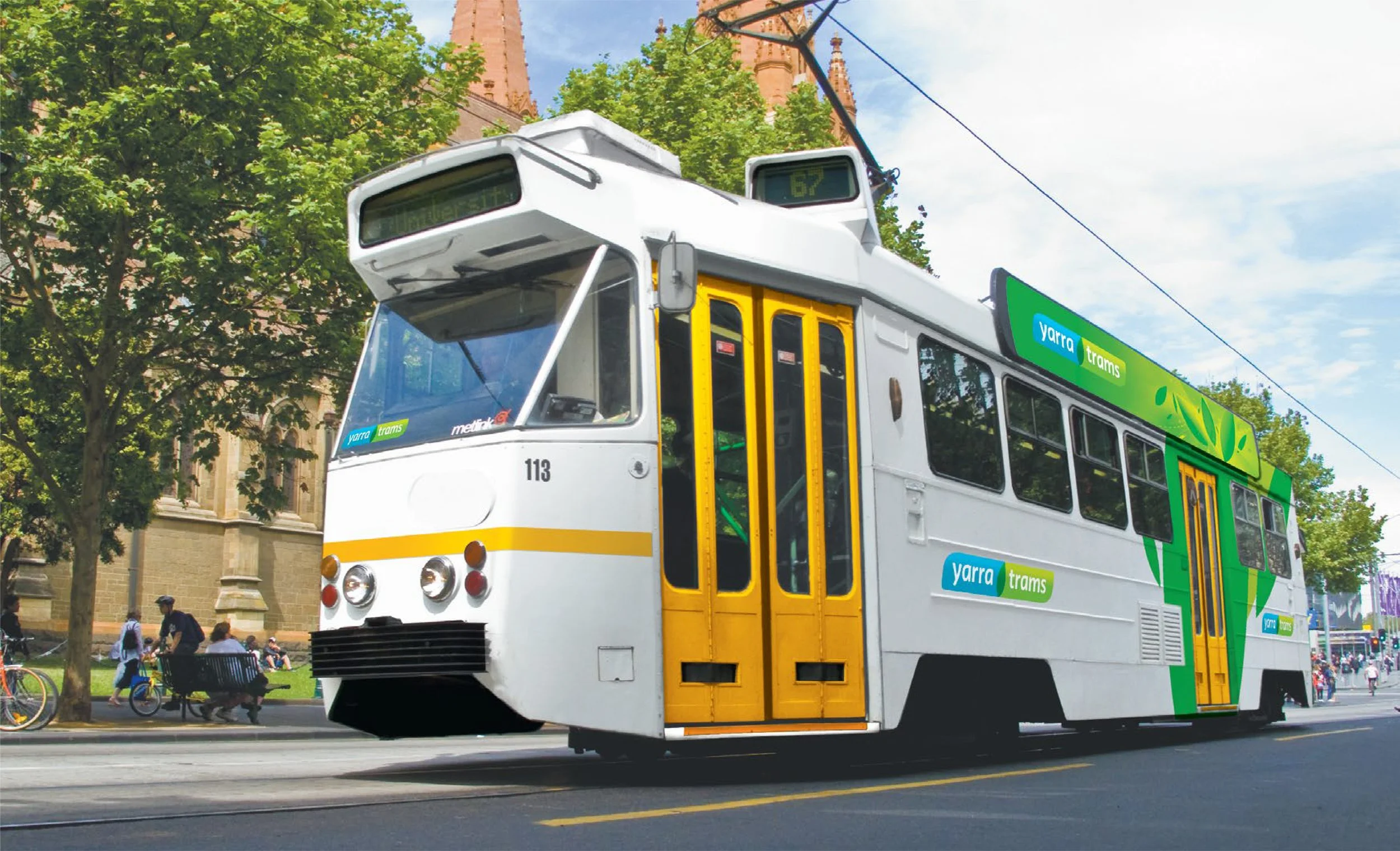 A tram with a green and white livery marked 'Yarra Trams' is traveling along a city street with trees and historic buildings in the background. People are walking and riding bicycles on the sidewalk.