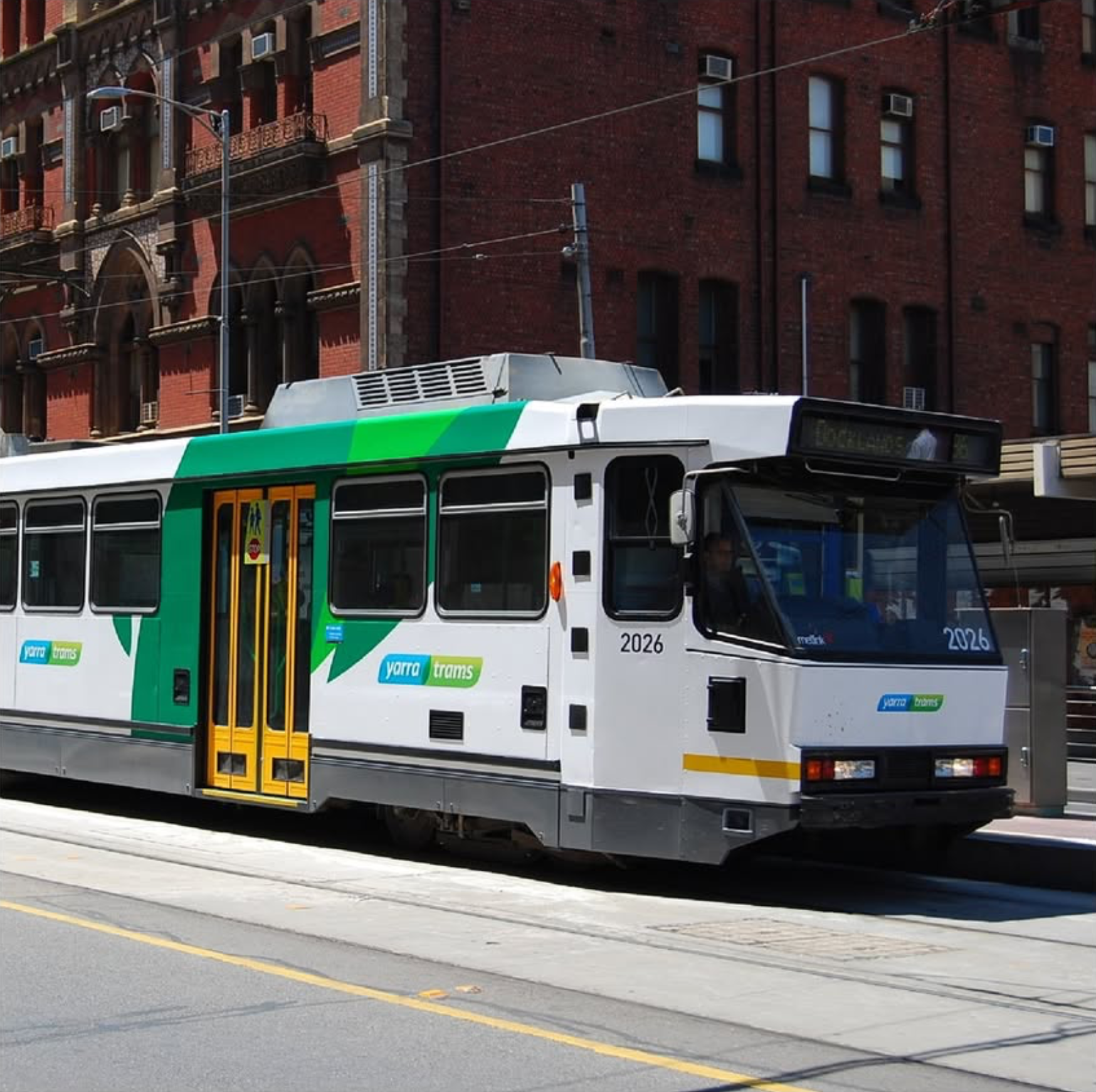 A white and green tram with the words 'Yarra Trams' on it is moving along a city street with yellow lane markings. In the background, there is a red brick building with multiple windows and some city electrical wires above.
