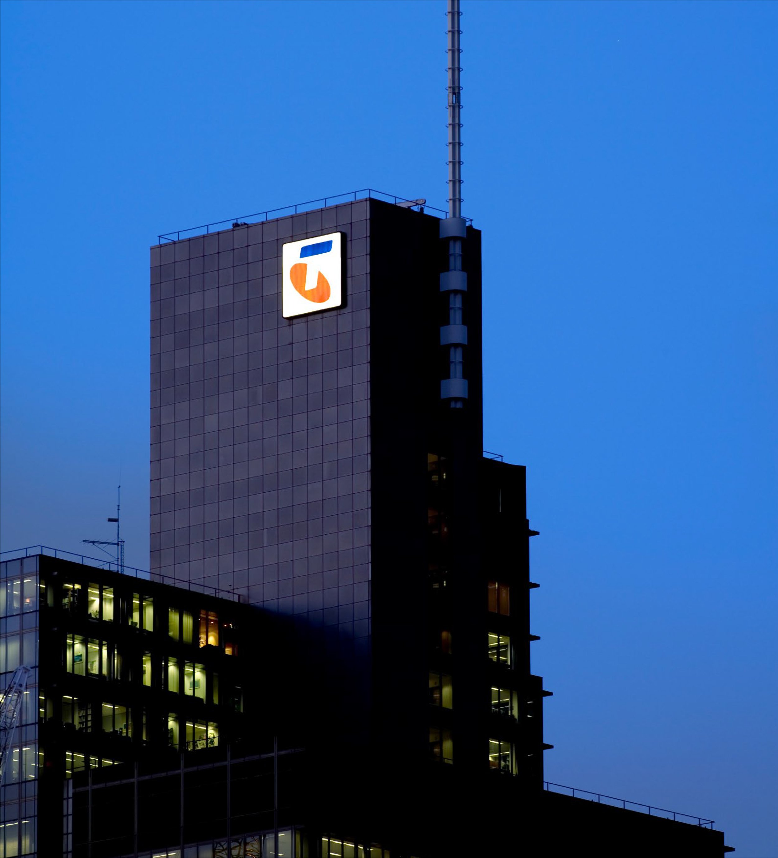 A tall modern building at dusk with illuminated windows and a bright logo on the facade, against a clear blue sky.
