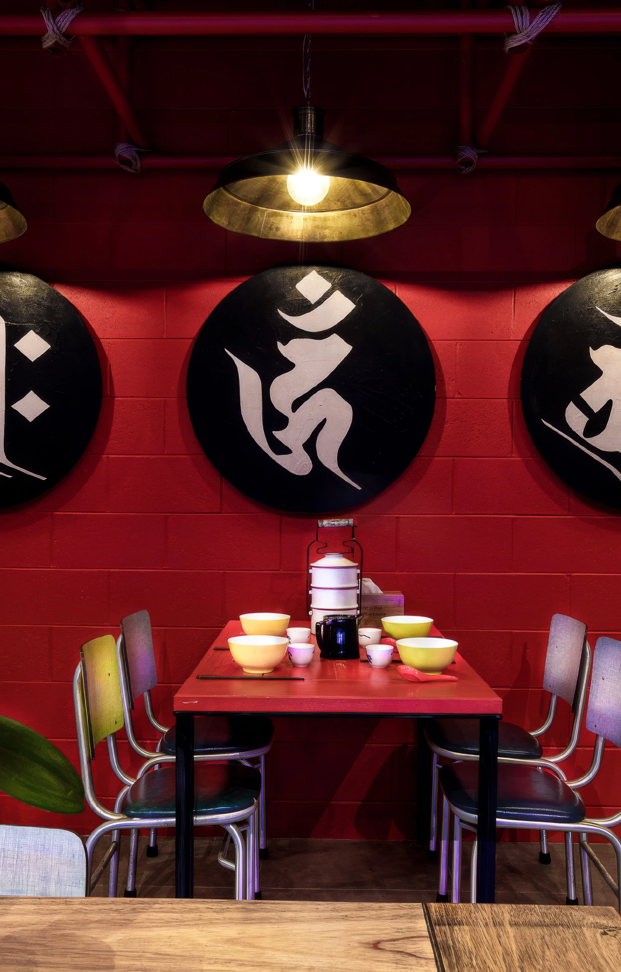 A dining table set with yellow and white bowls, cups, and a black teapot, with colorful chairs around it, against a red wall decorated with black circular artwork featuring white abstract designs. A hanging light illuminates the scene.