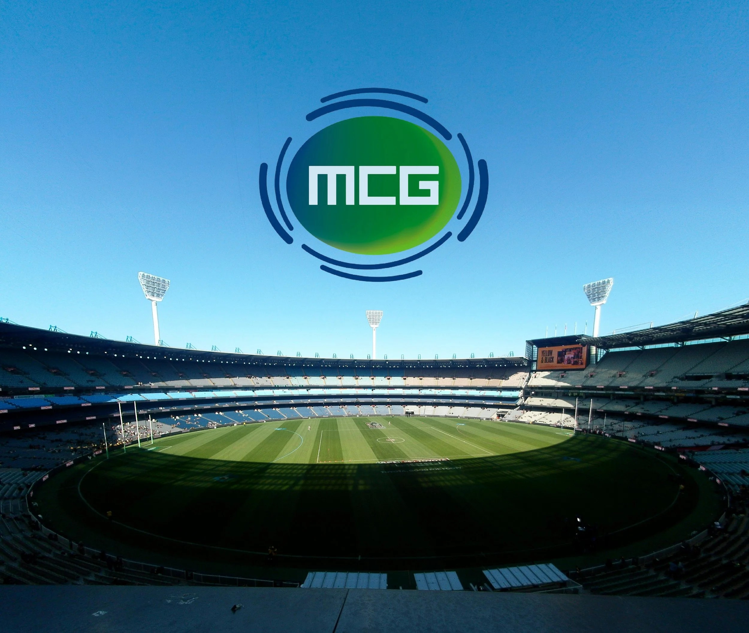 Empty stadium with a green football field and blue seats, under a clear blue sky, with large lighting structures and a scoreboard.