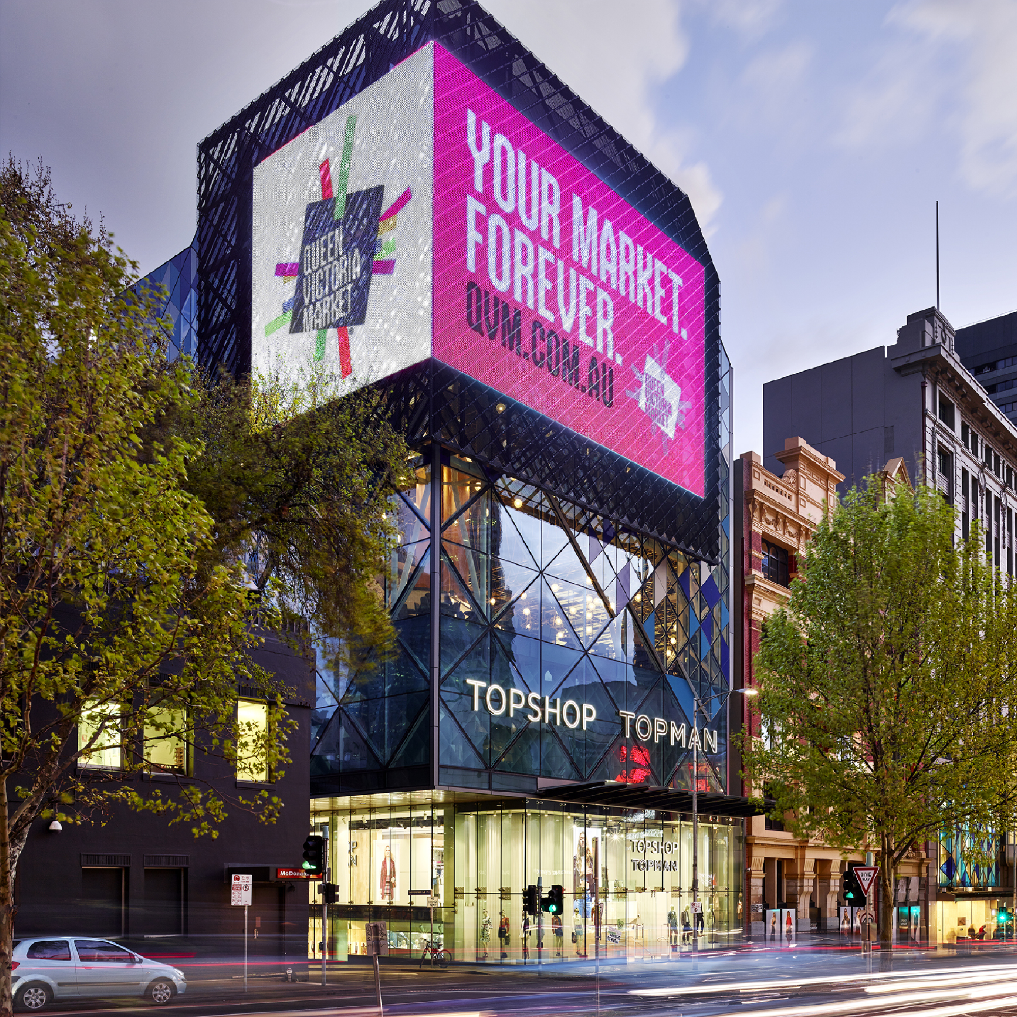 A modern multi-story building with a glass exterior displaying a large digital billboard. The billboard shows an advertisement for Topshop and Topman, featuring mannequins wearing fashionable clothing. There are green traffic lights, a car passing by