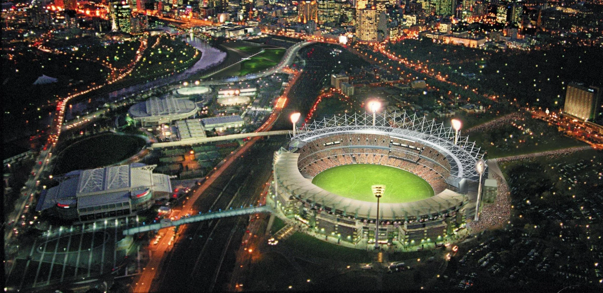 Night aerial view of a large football stadium filled with spectators, illuminated with bright lights, surrounded by city streets and buildings with glowing lights in the background.