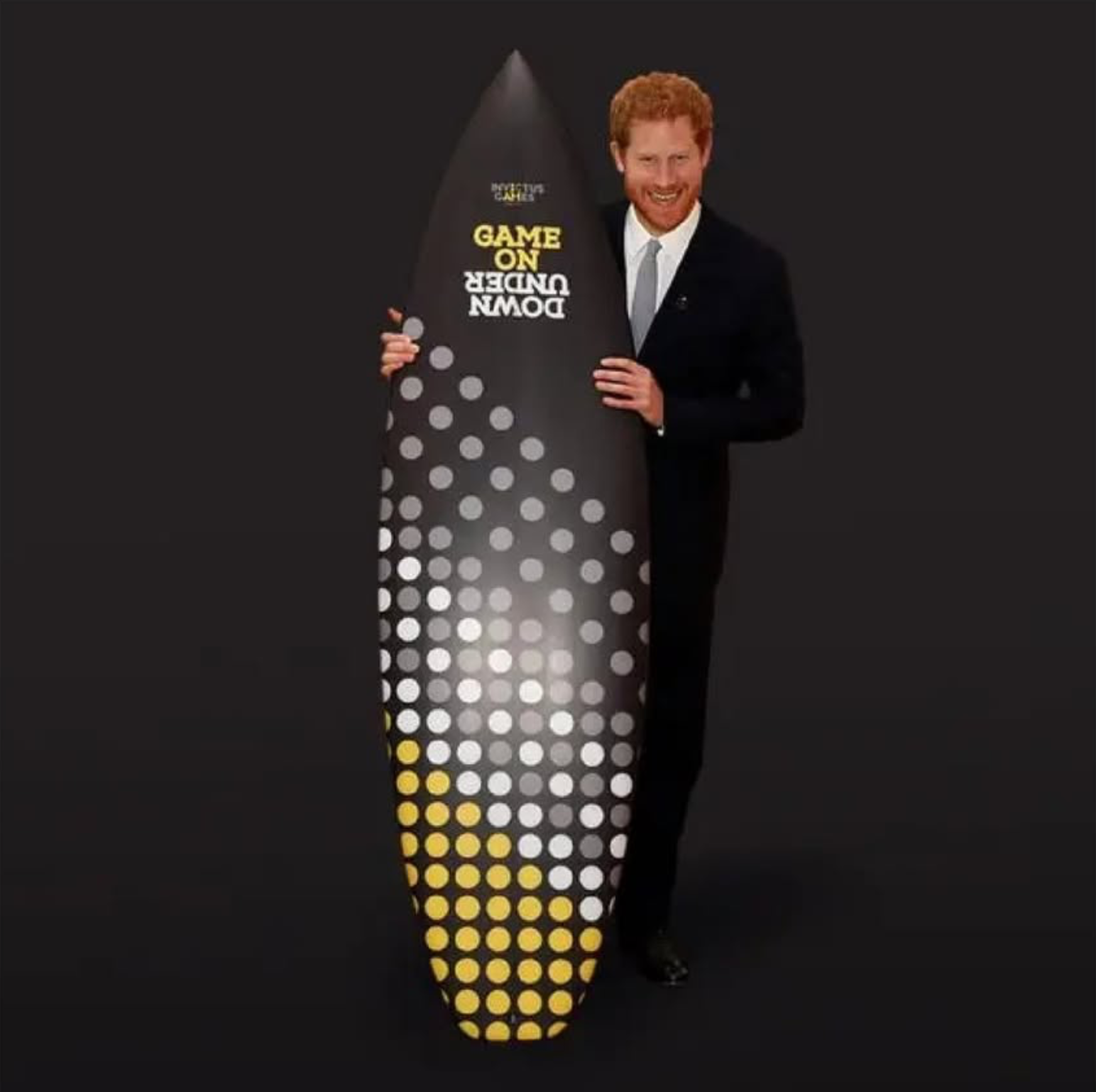 Prince Harry in a suit holding a large award shaped like a rocket with the words 'Down Under' and 'Game On' written on it in yellow and white text. The award is decorated with white and yellow dots, and the background is black.