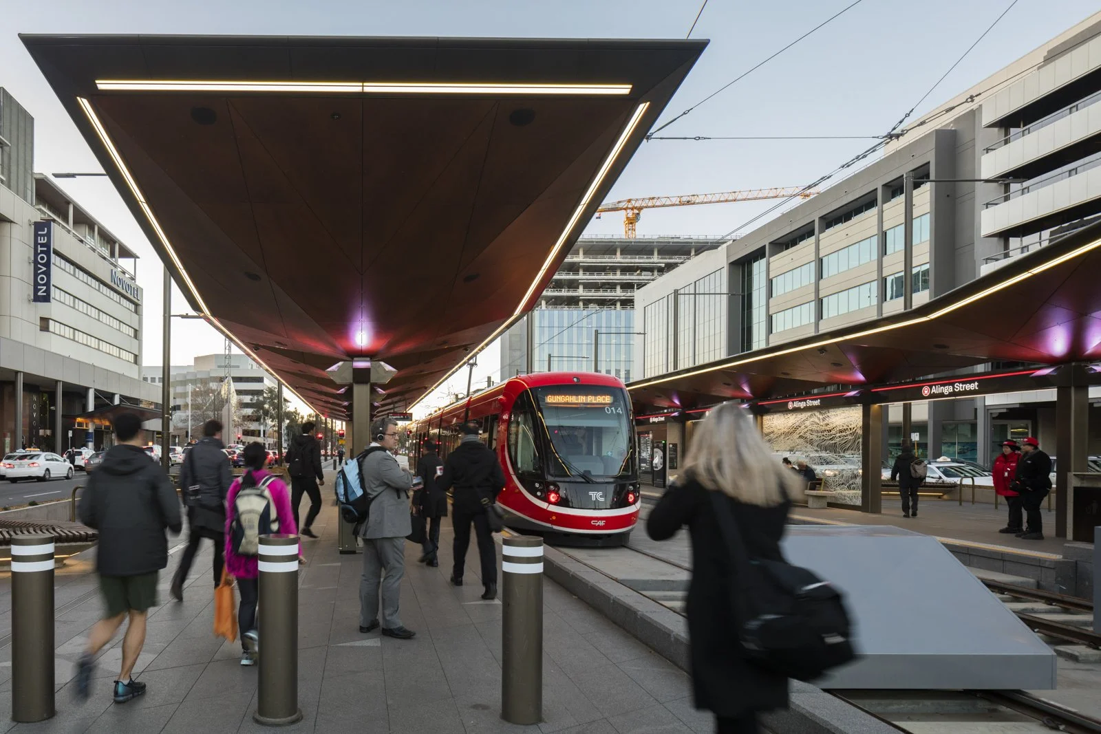 People walking and waiting at a modern city train station with a red train on the tracks, surrounded by buildings and a clear sky.