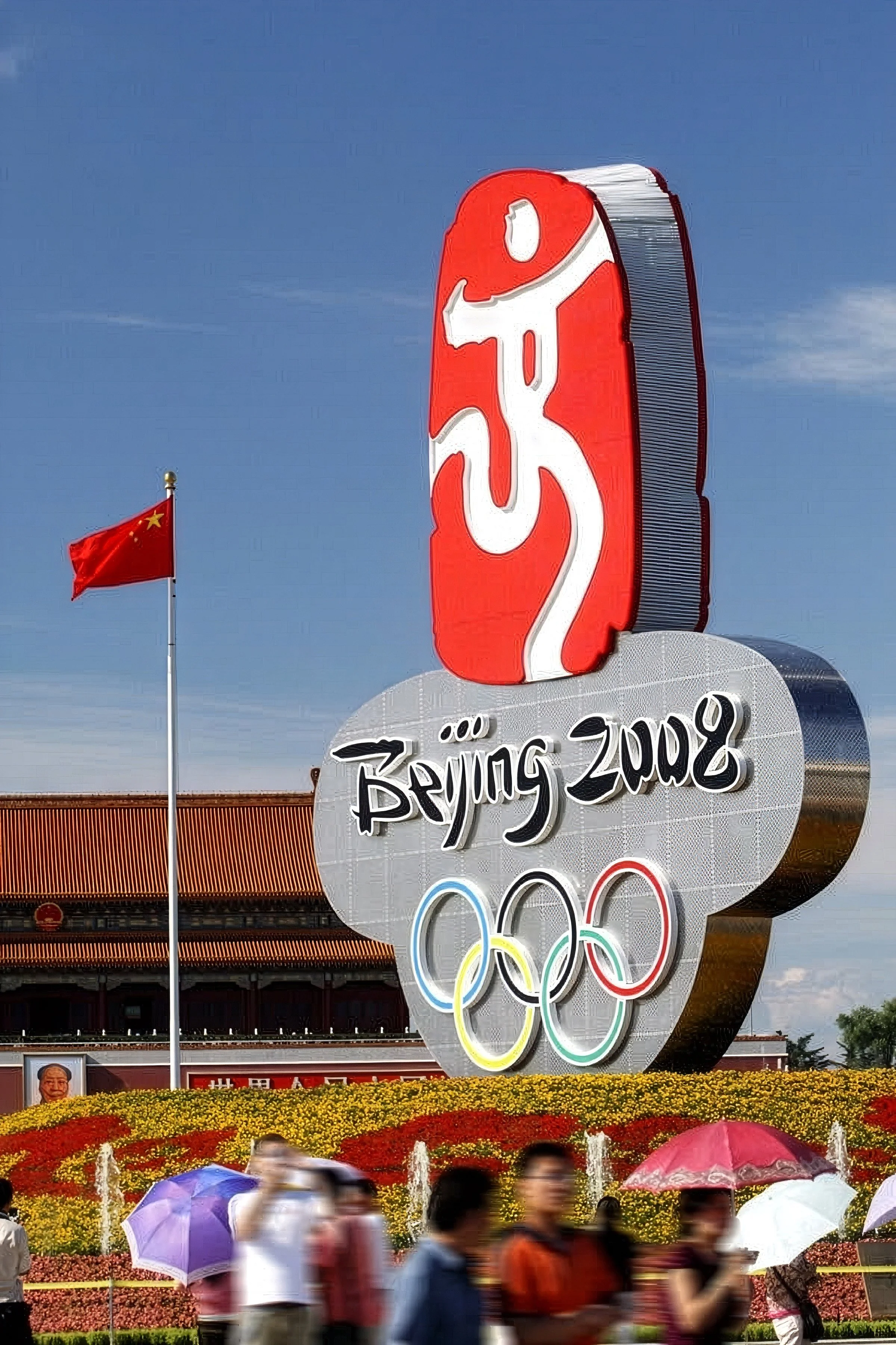 Large sign displaying the Beijing 2008 Olympic Games logo with the Olympic rings, alongside a Chinese flag on a pole and a crowd of people with umbrellas in a park area.