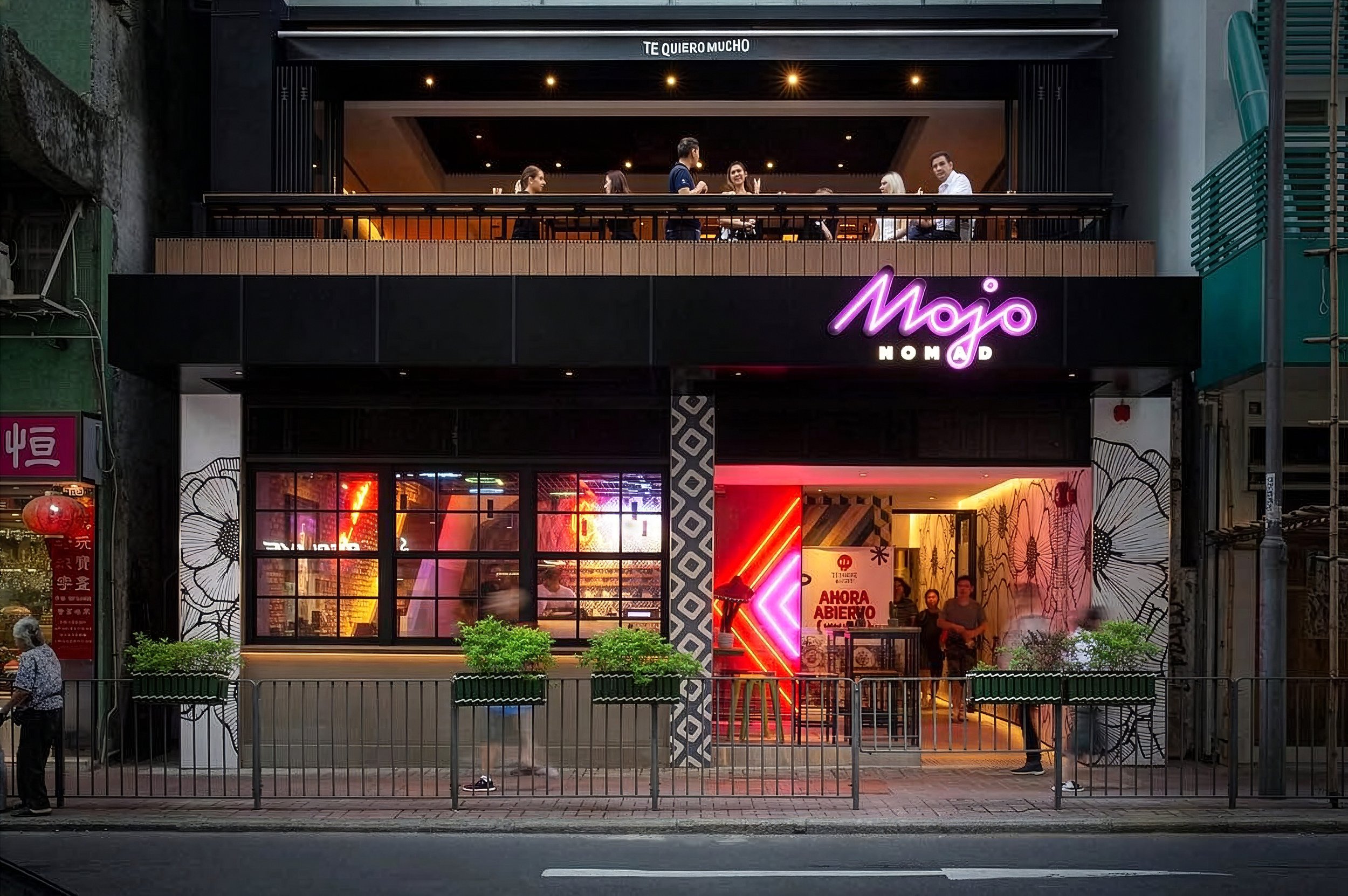 Night view of a two-story restaurant or cafe called 'Mojó' with neon signage. The upper floor has people dining and socializing, with warm interior lighting. The lower floor features large windows, colorful neon lights, and a sign in Spanish saying 'AHORA ABIERTO'. There is a sidewalk with a metal fence, and a few pedestrians are seen in front of the building.