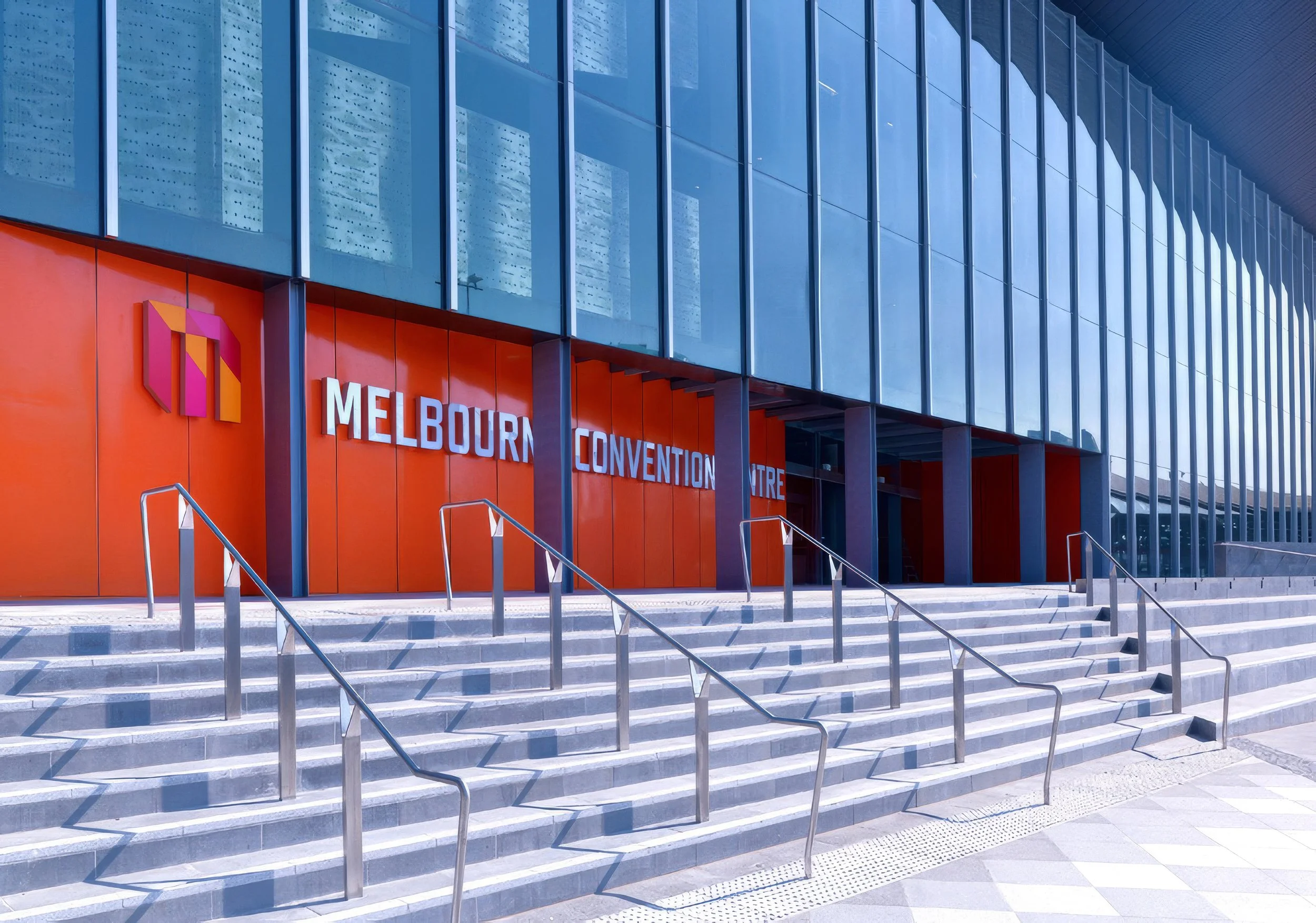 Exterior view of the Melbourne Convention Centre with orange walls, large glass windows, and stone stairs with metal railings.