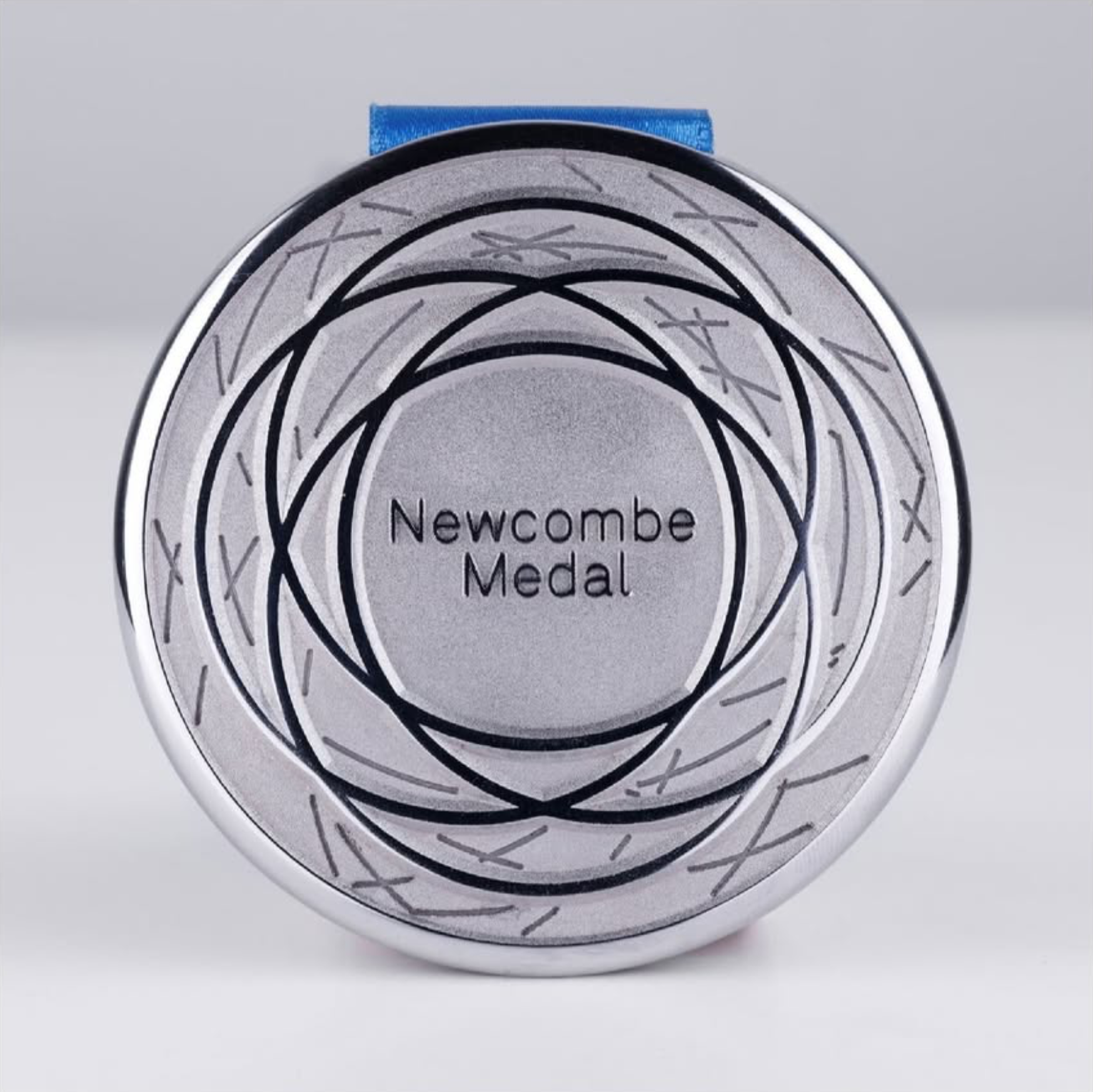A round, silver-colored medal with black and gray concentric and overlapping circular patterns. The center of the medal displays the text 'Newcombe Medal'.