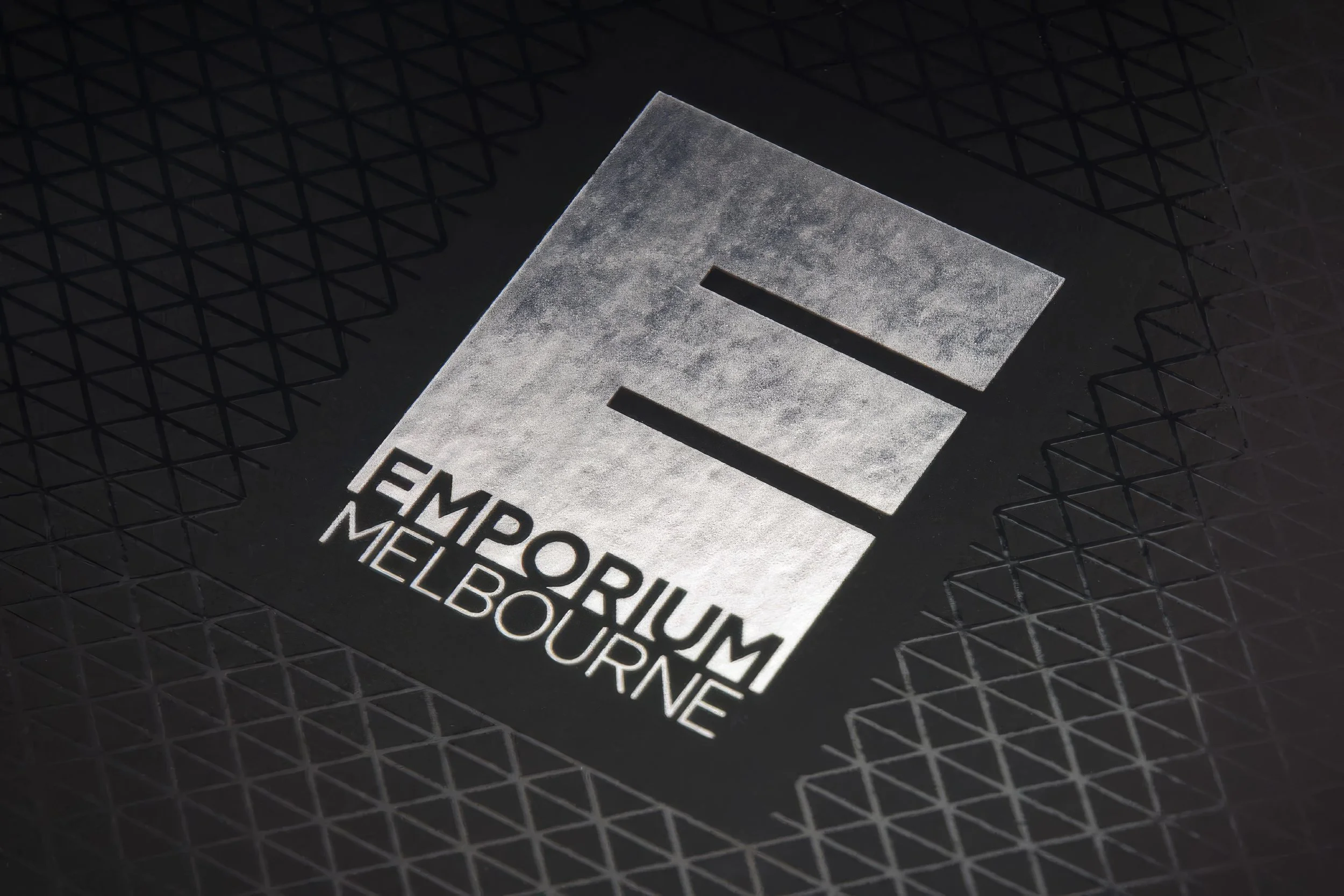 Close-up of a metallic sign on a black textured background that reads 'EMPORIUM MELBOURNE' with three horizontal bars above the text.