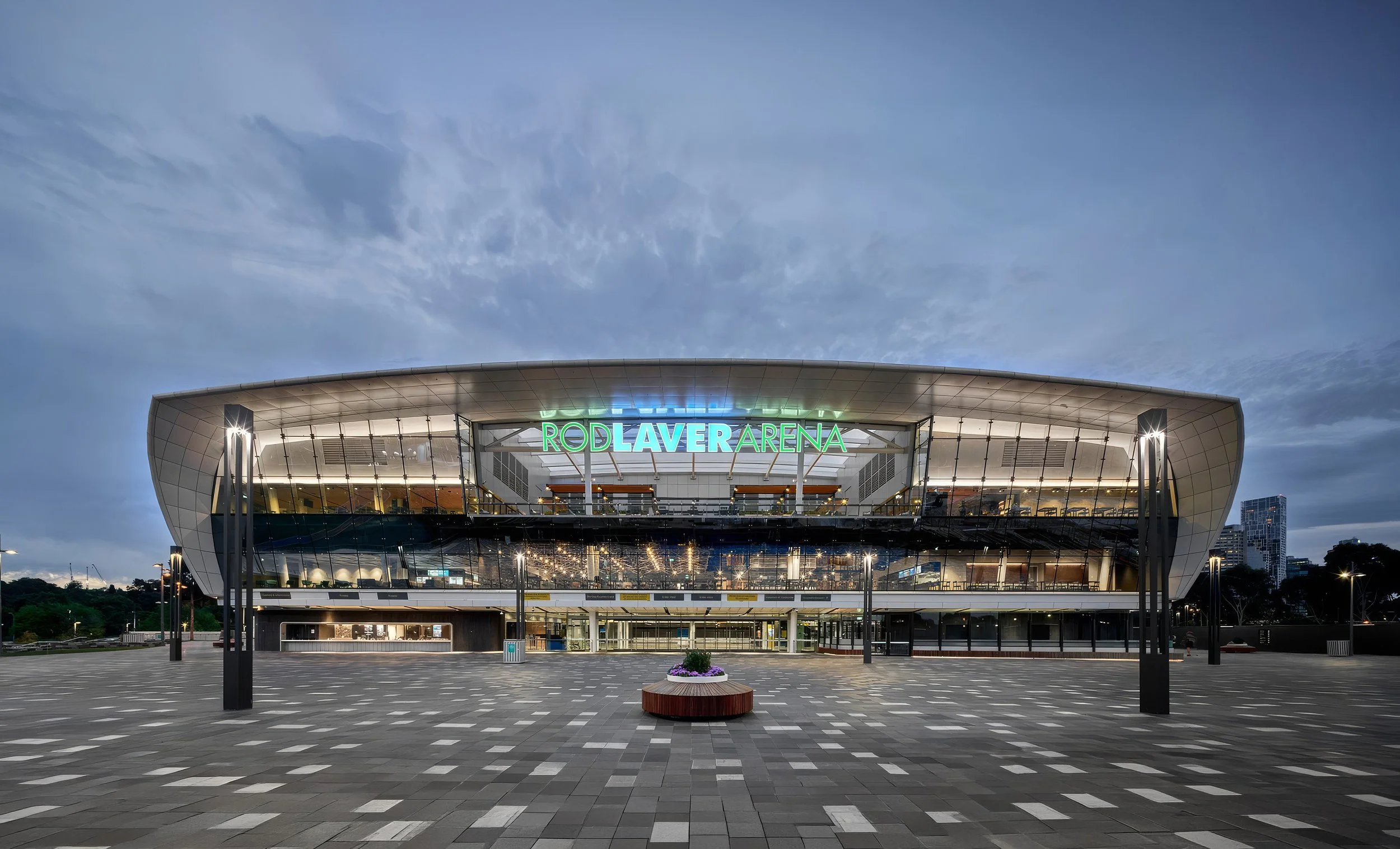 Exterior of Rod Laver Arena during dusk with illuminated sign, glass facade, and checkered pavement.