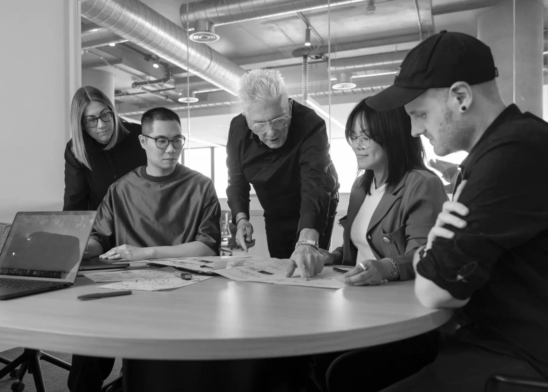 Group of five people gathered around a table, discussing and looking at papers, with a laptop open in front of them, in a modern office space.