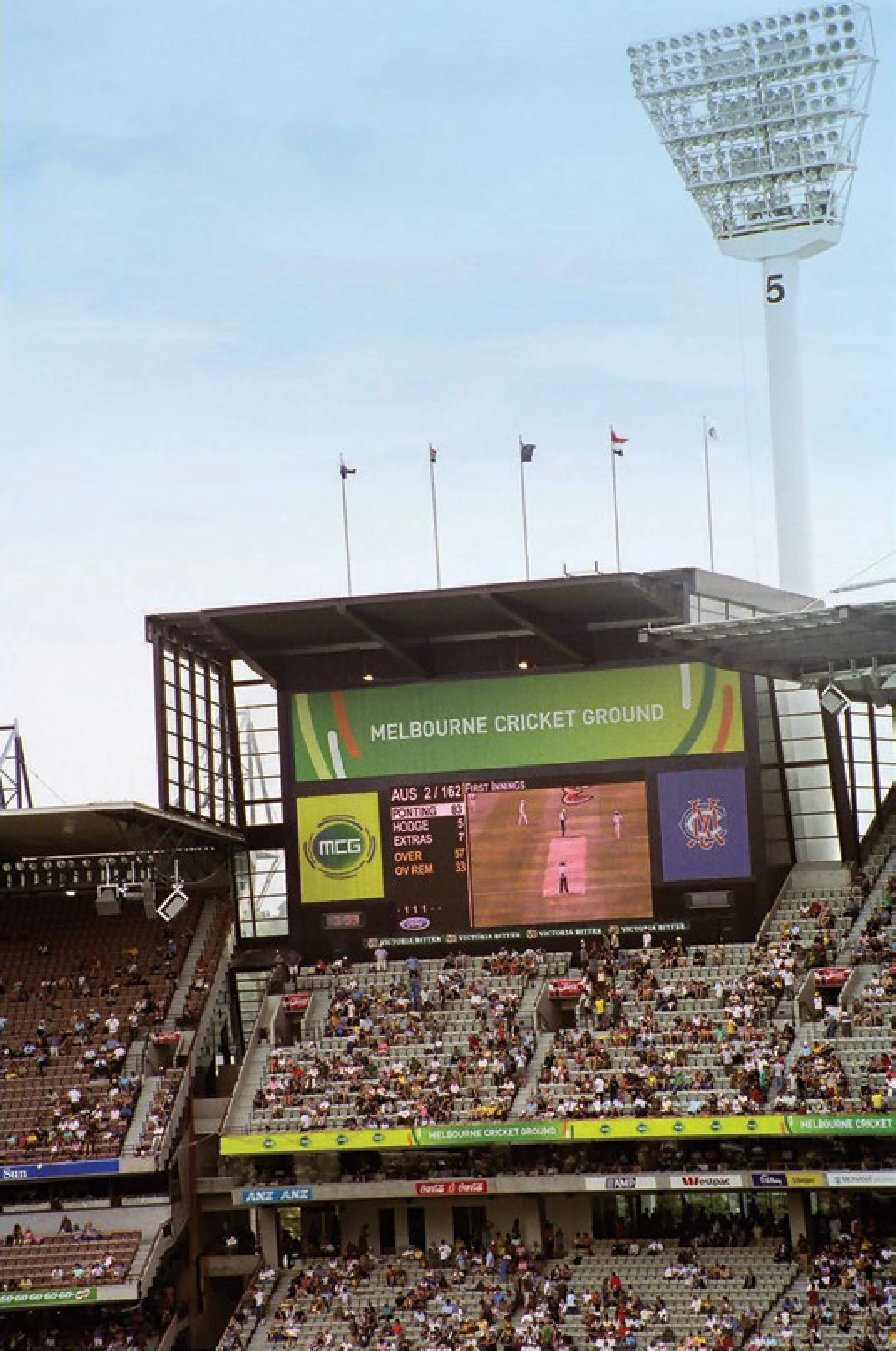A large outdoor stadium filled with spectators watching a cricket match at Melbourne Cricket Ground, with a scoreboard displaying scores and flags flying on poles. A tall floodlight tower is visible in the background.