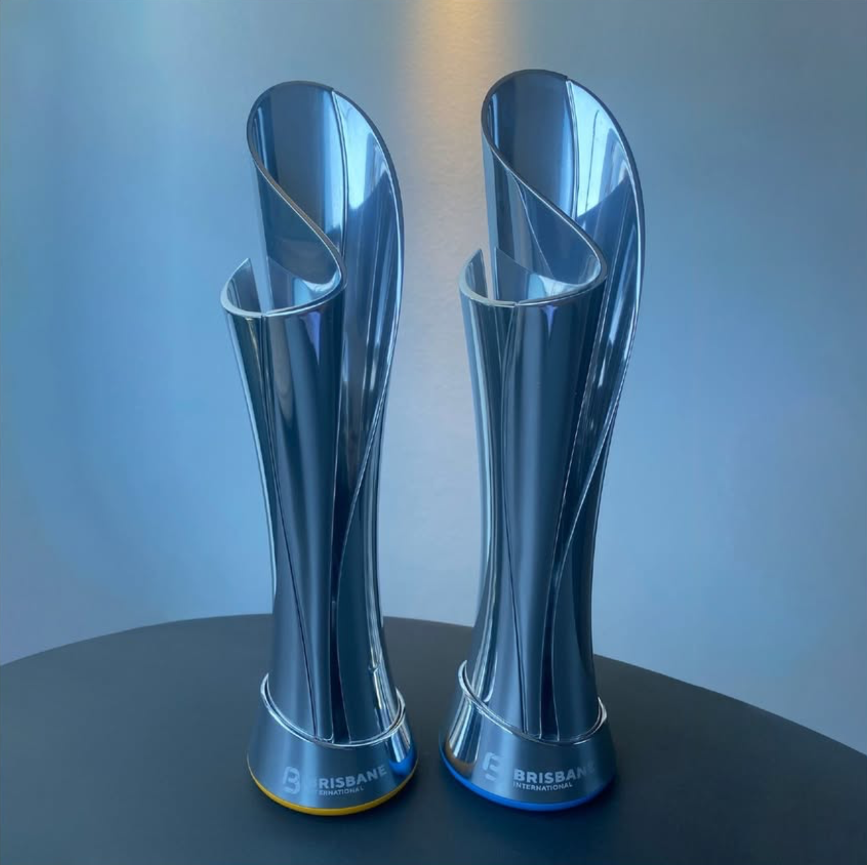 Two metallic silver, blue, and yellow souvenir cups with a swirl design, displaying the word 'BRISBANE' and 'INTERNATIONAL', placed on a dark surface against a neutral background.