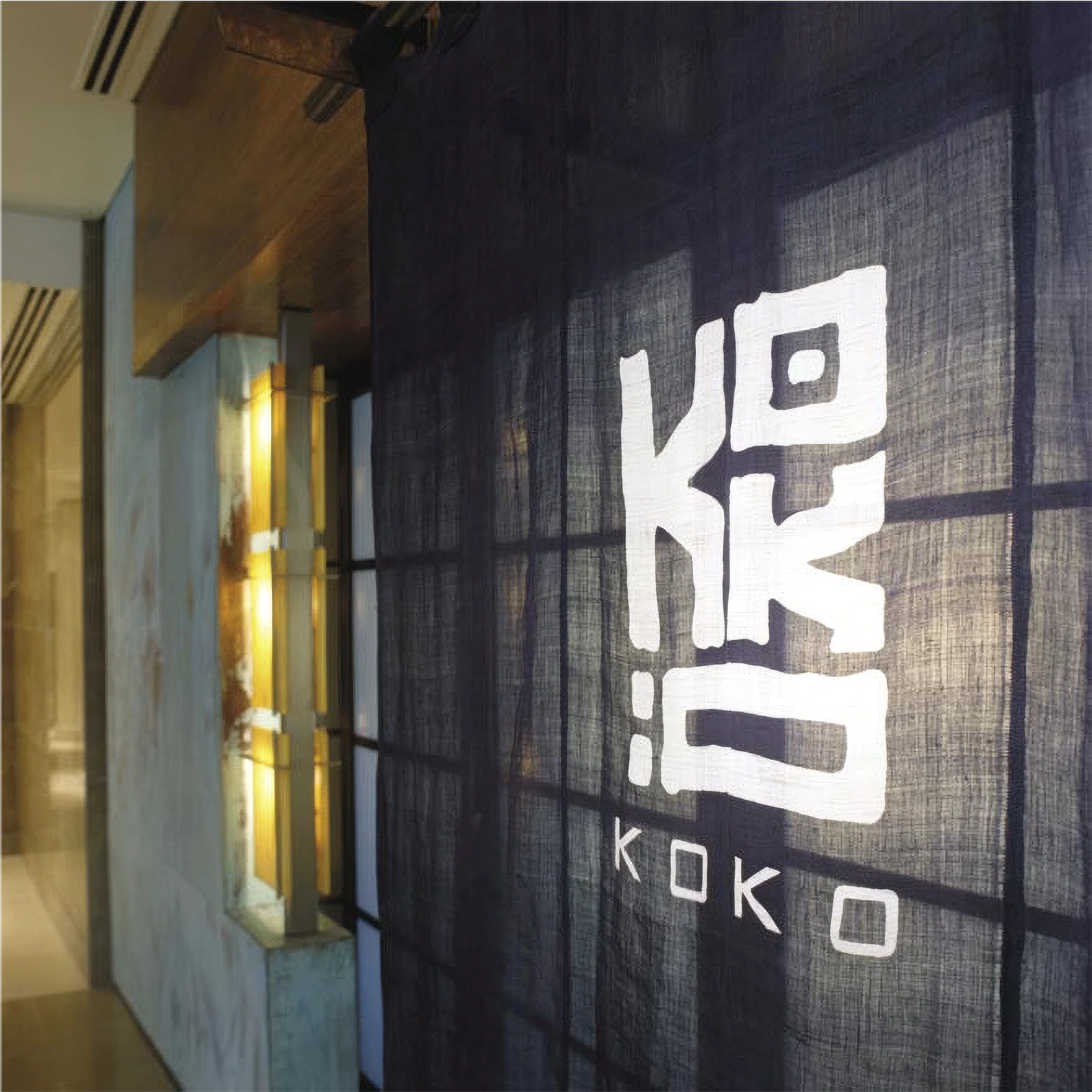 Interior of a modern building with a black wall featuring large white block letters spelling 'KOKO'. Yellow lighting accents and wooden ceiling panels. An air conditioning vent and some furniture are visible.