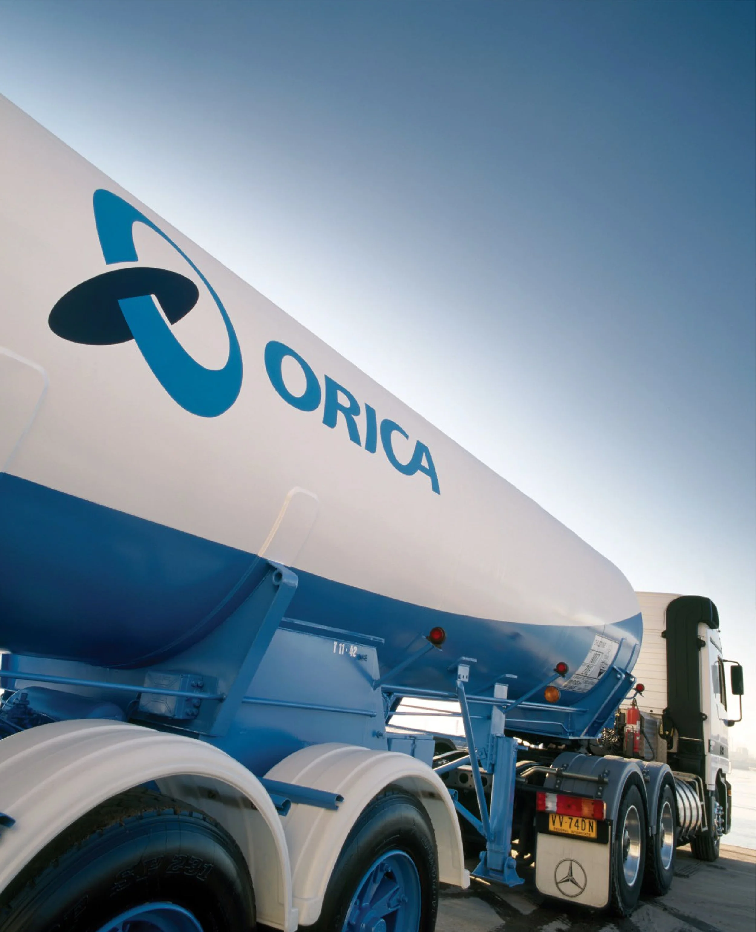 Side view of a large truck with multiple blue and white tanks and wheels, part of the Mercedes-Benz logo visible on the mud flap, parked outdoors under clear sky.
